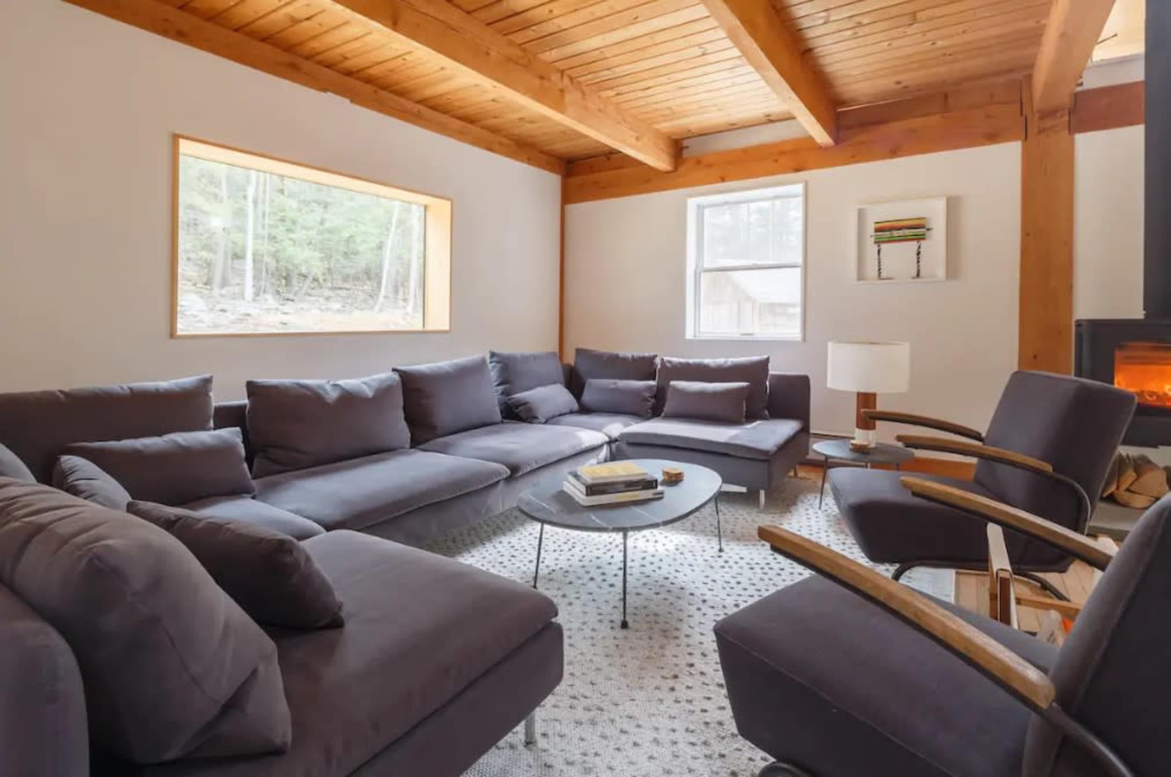 The image shows a modern living room with a large sectional sofa, two wooden armchairs, a coffee table, and windows allowing natural light to enter.