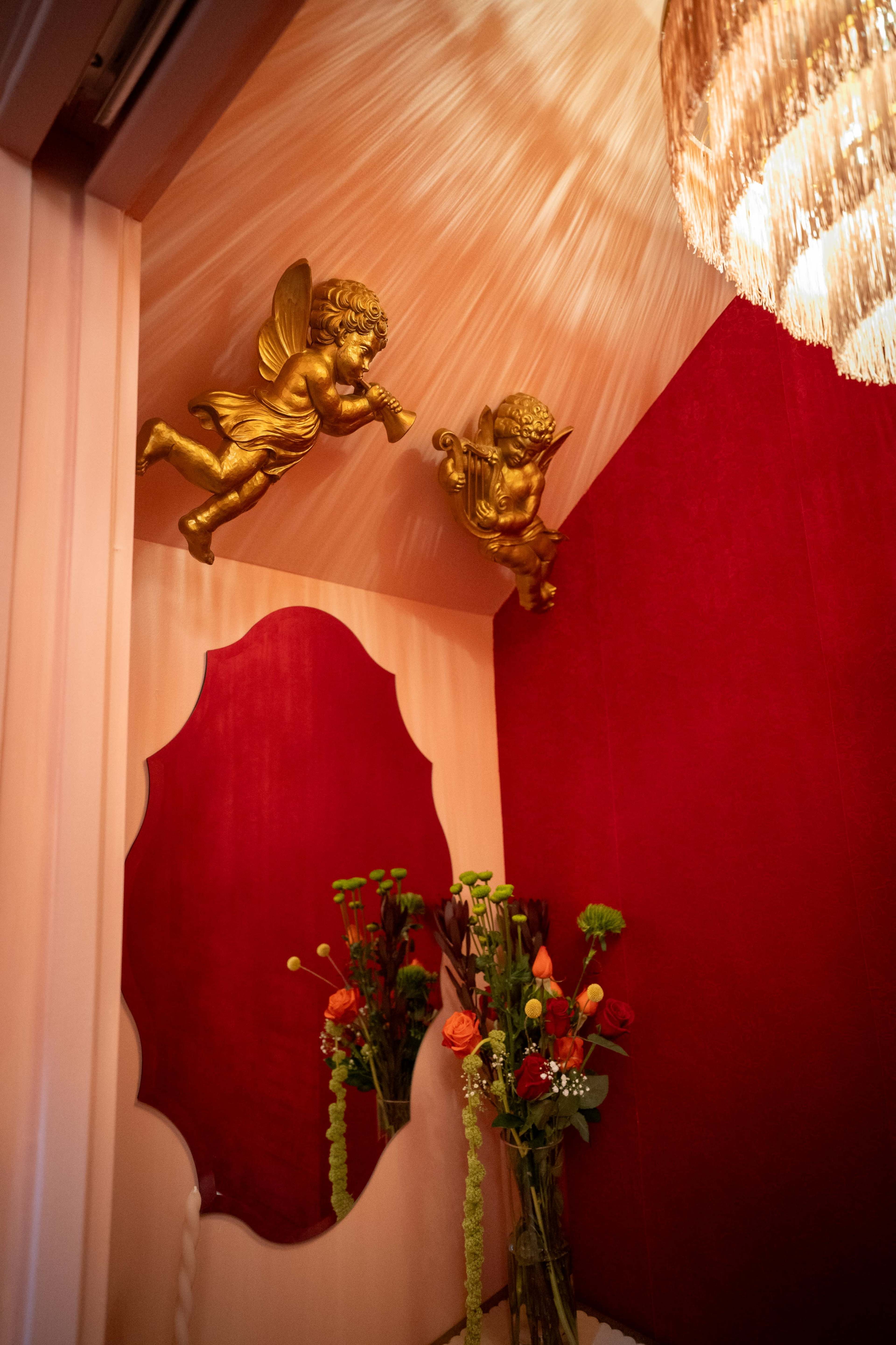 The image shows a corner of a room with red walls, golden cherub figurines on the ceiling, a decorative mirror, and a bouquet of flowers on the floor.