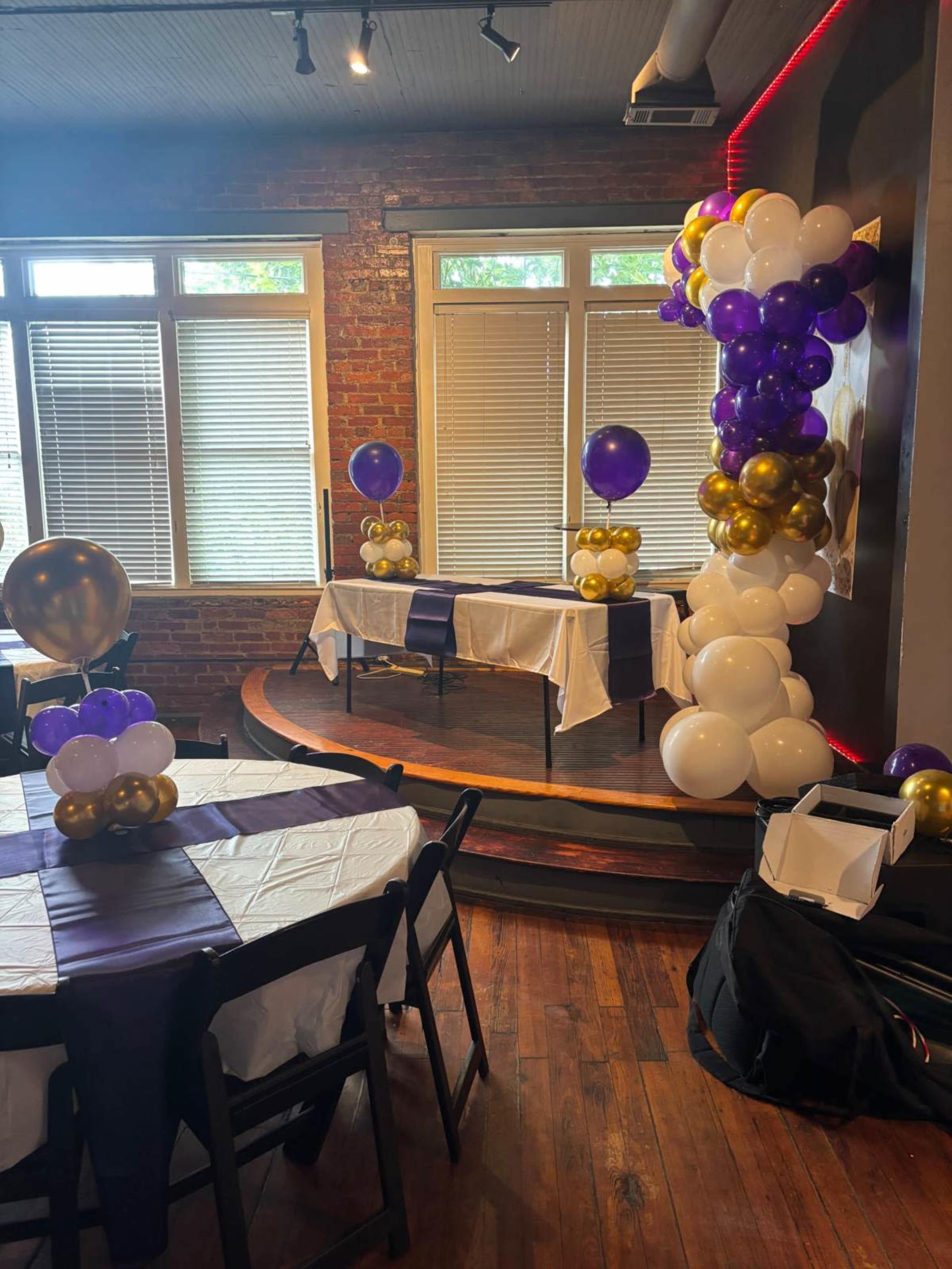 The image shows a small event space with a wooden stage decorated with balloons in purple, gold, and white, and tables set with white linens and dark purple accents.