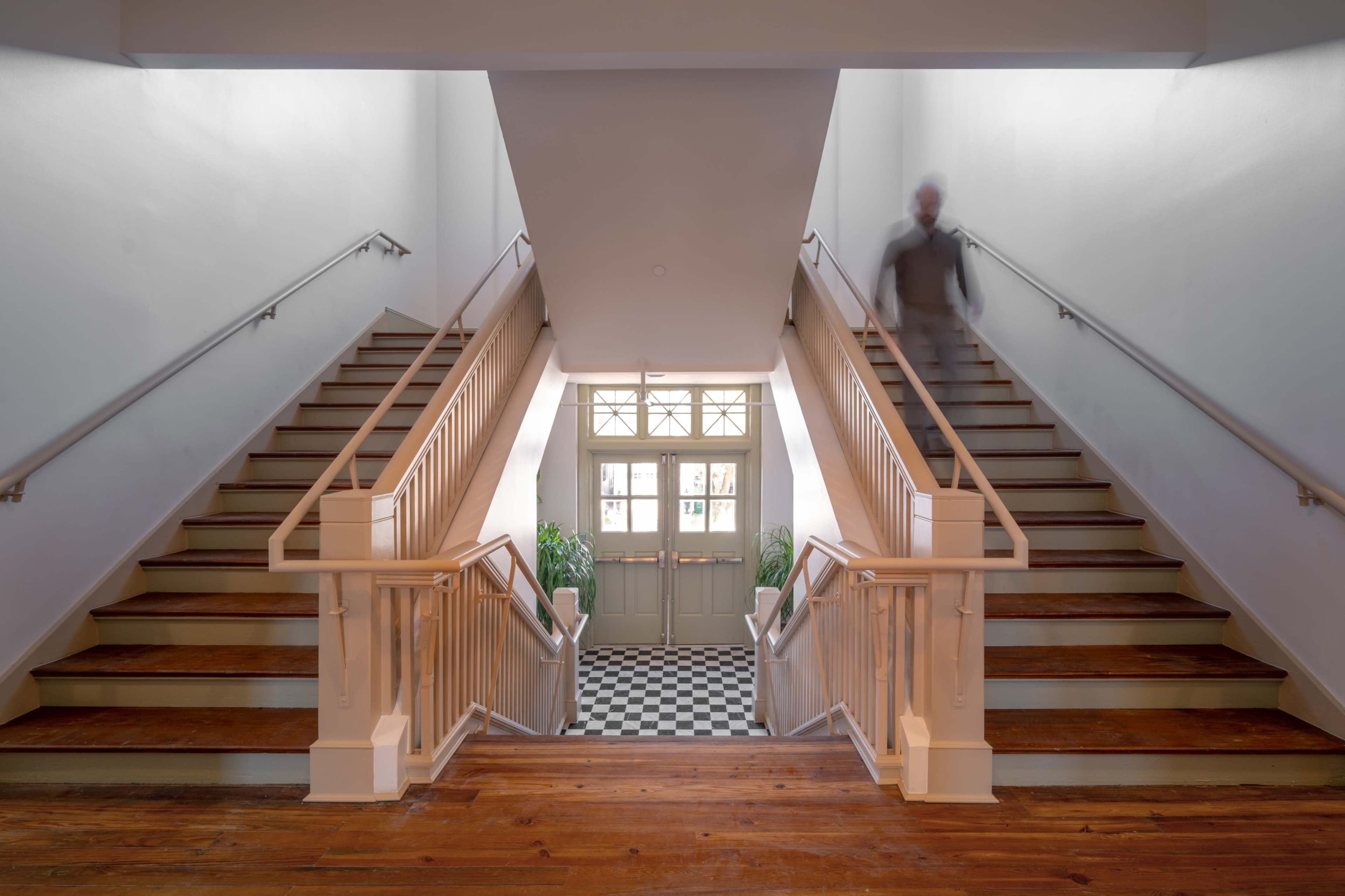The image shows a symmetrical staircase with wooden steps and white railings leading to a pair of doors at the center, with a checkered floor visible below.