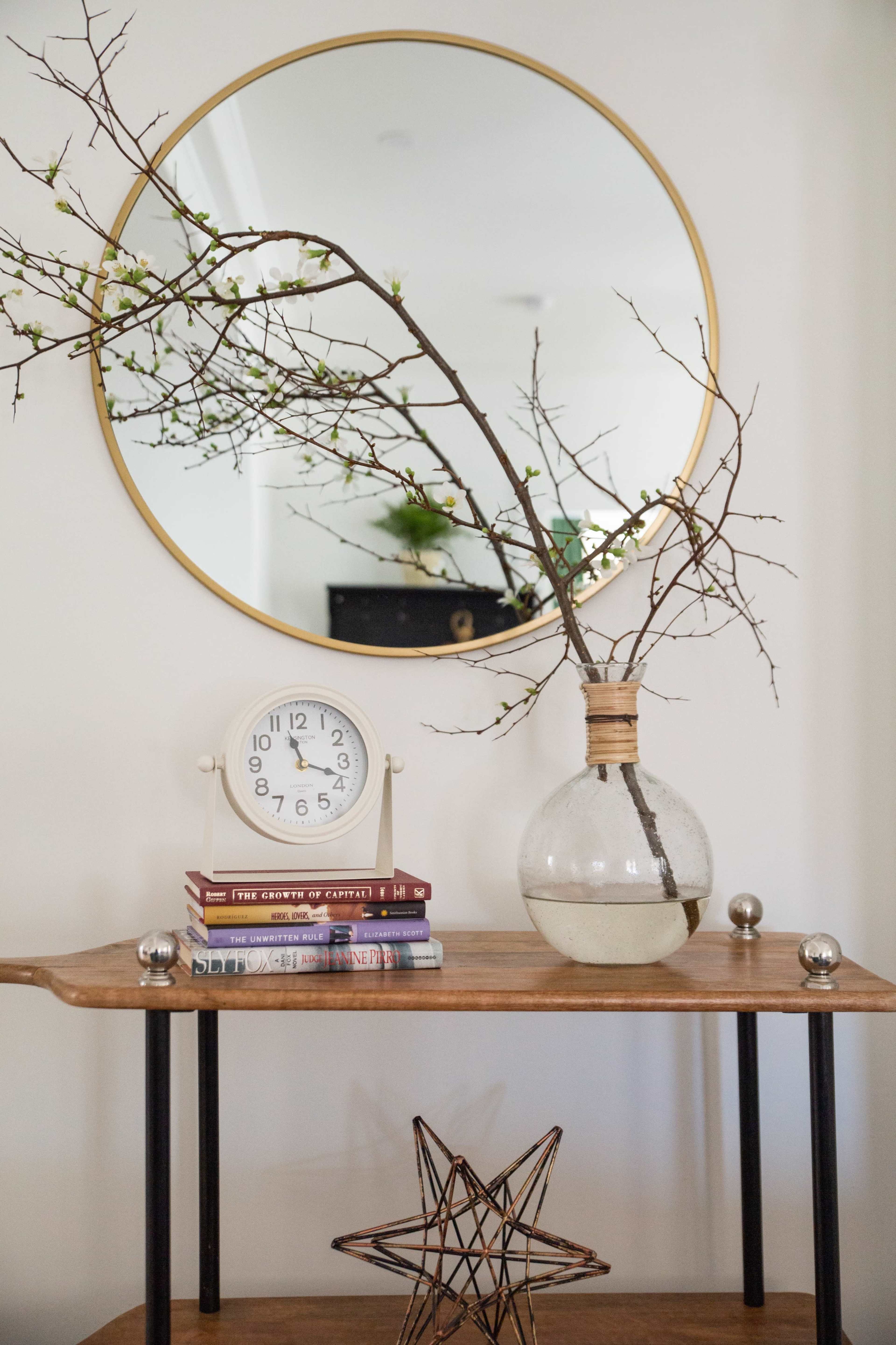 A wooden table displays a round mirror, a glass vase with a bare branch, a clock, and several stacked books.