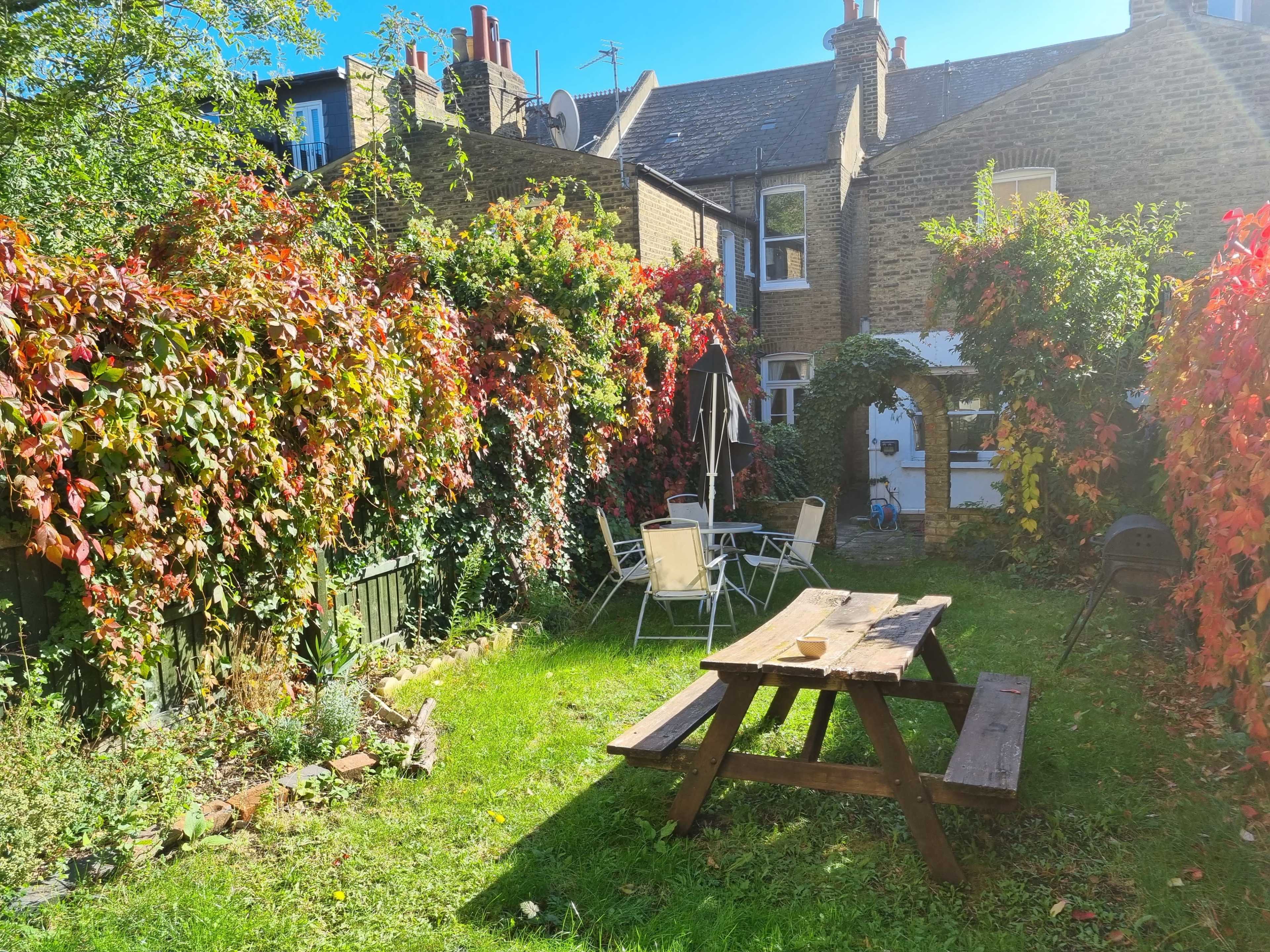 A garden features a wooden picnic table surrounded by colorful foliage and a small seating area near a white door in a brick house.