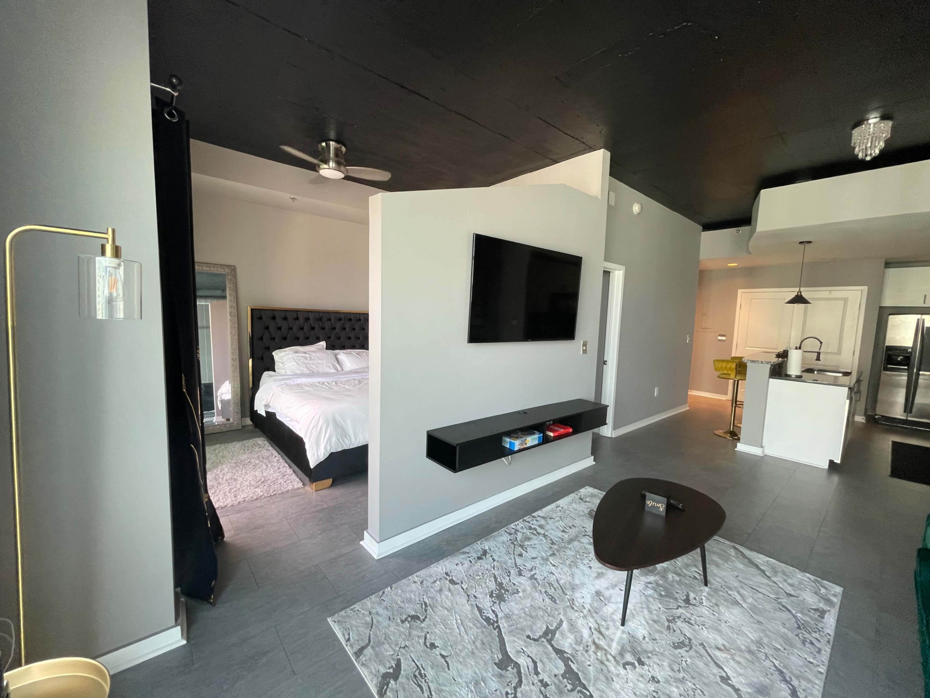 The image shows a modern, open-concept living space featuring a bedroom area with a bed, a living area with a coffee table and television, and a kitchenette visible in the background.
