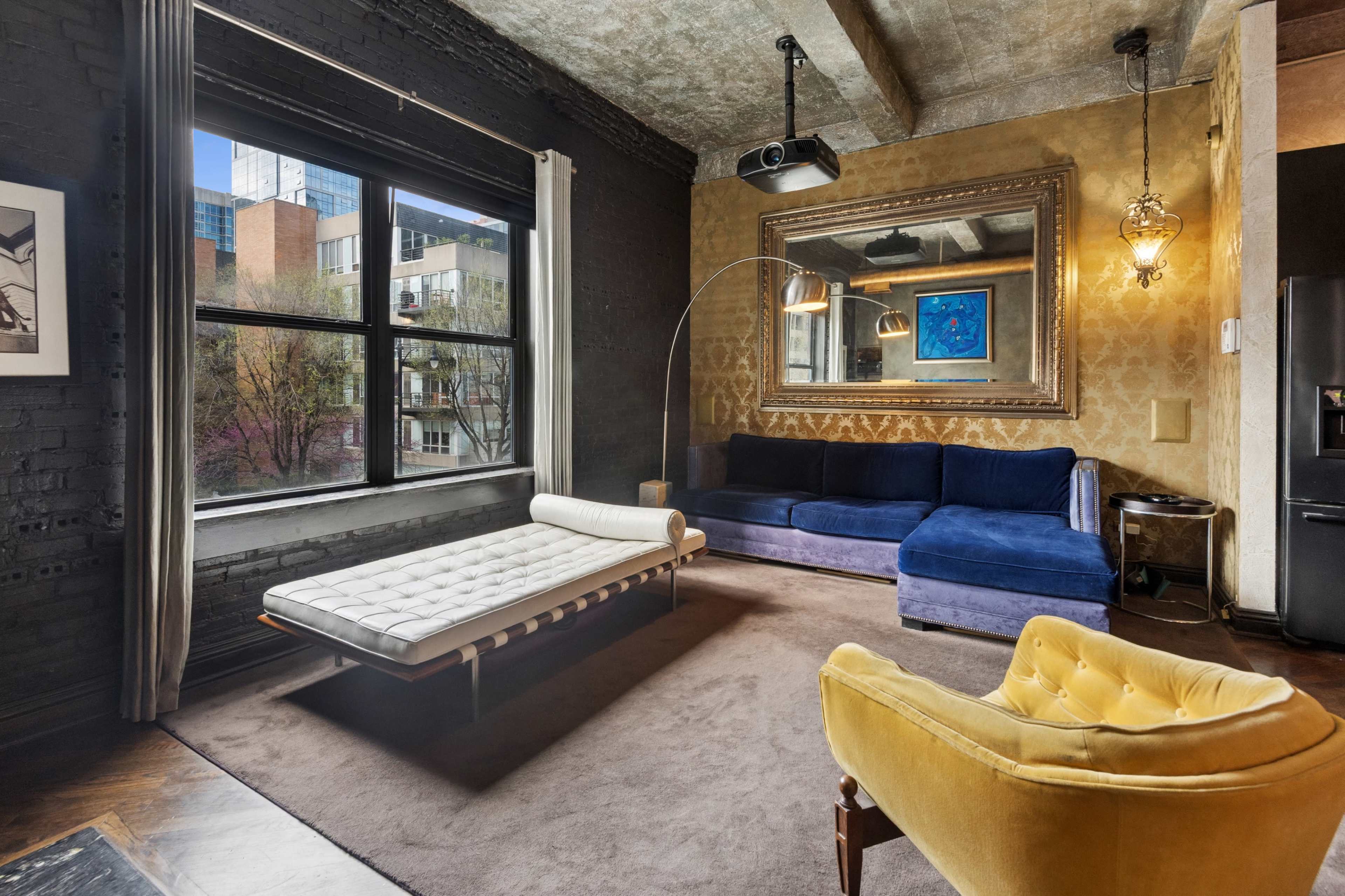 The image shows a stylish living room featuring a dark wall, a large window, a blue sofa, a yellow armchair, and a decorative wall mirror.