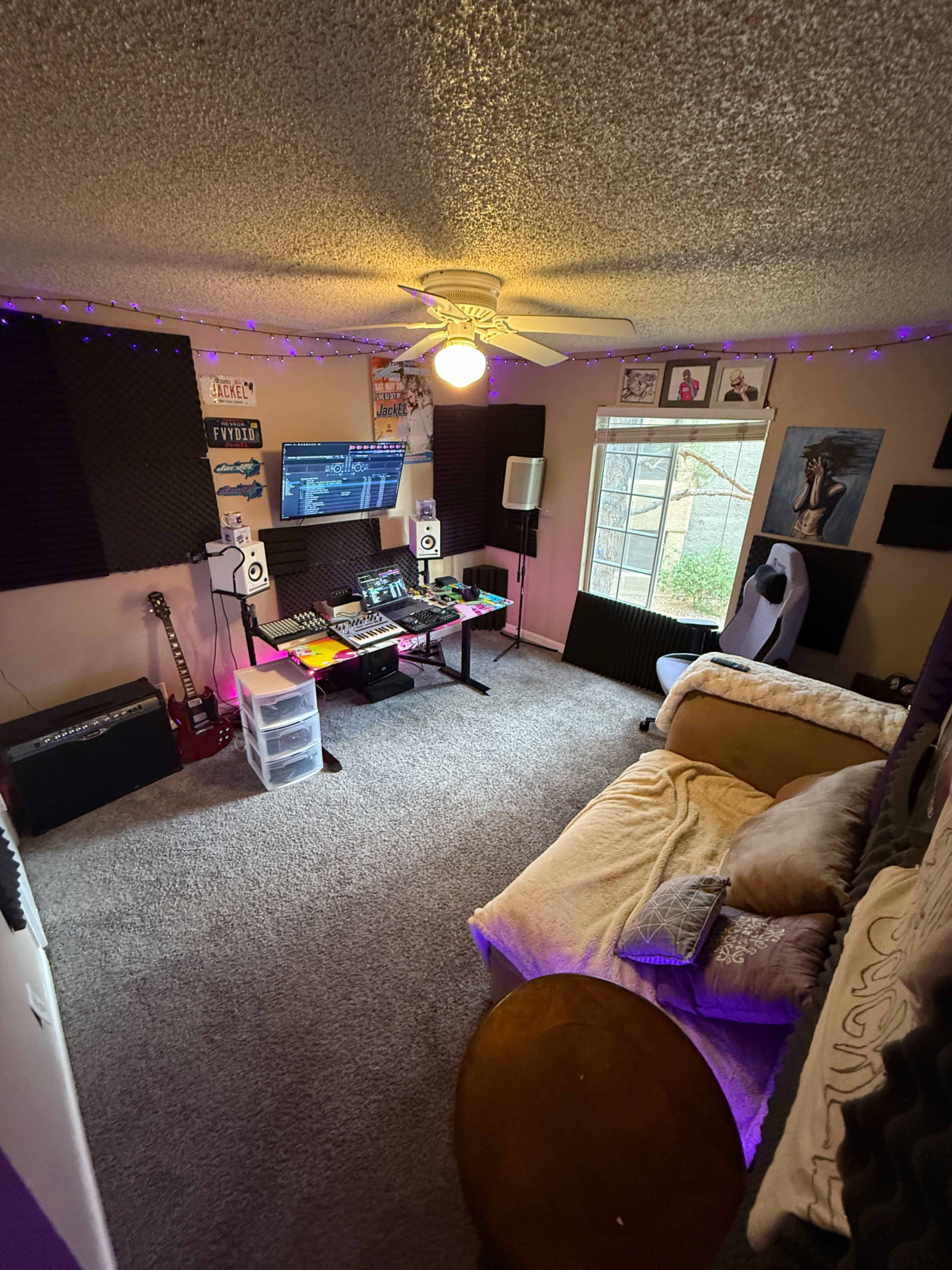 The image depicts a cozy music studio with soundproofing panels on the walls, a couch, a computer setup, and various musical instruments.