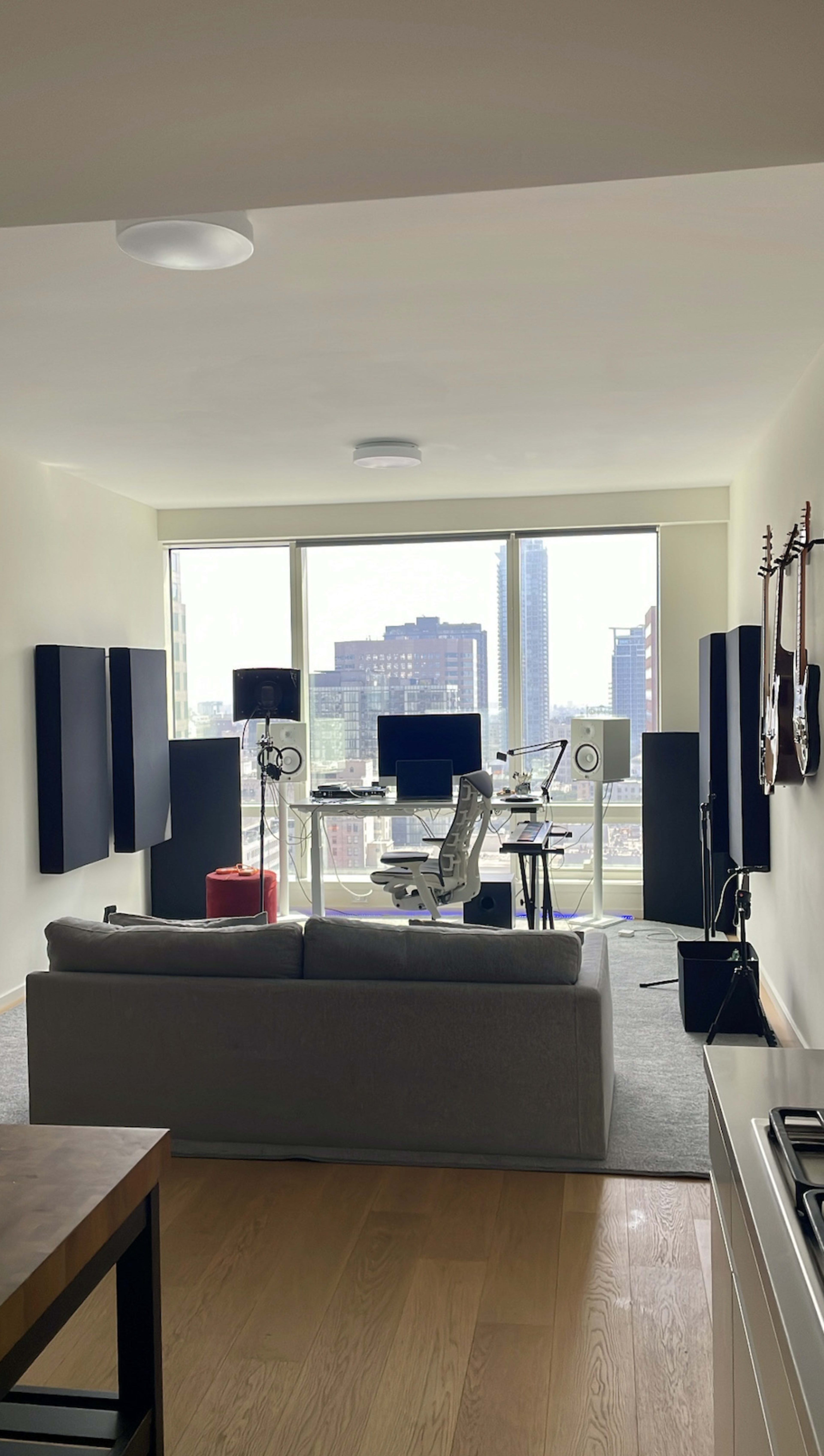 The image shows a modern living room with a gray sofa, a workstation equipped with electronic devices, and large windows offering a view of a cityscape.