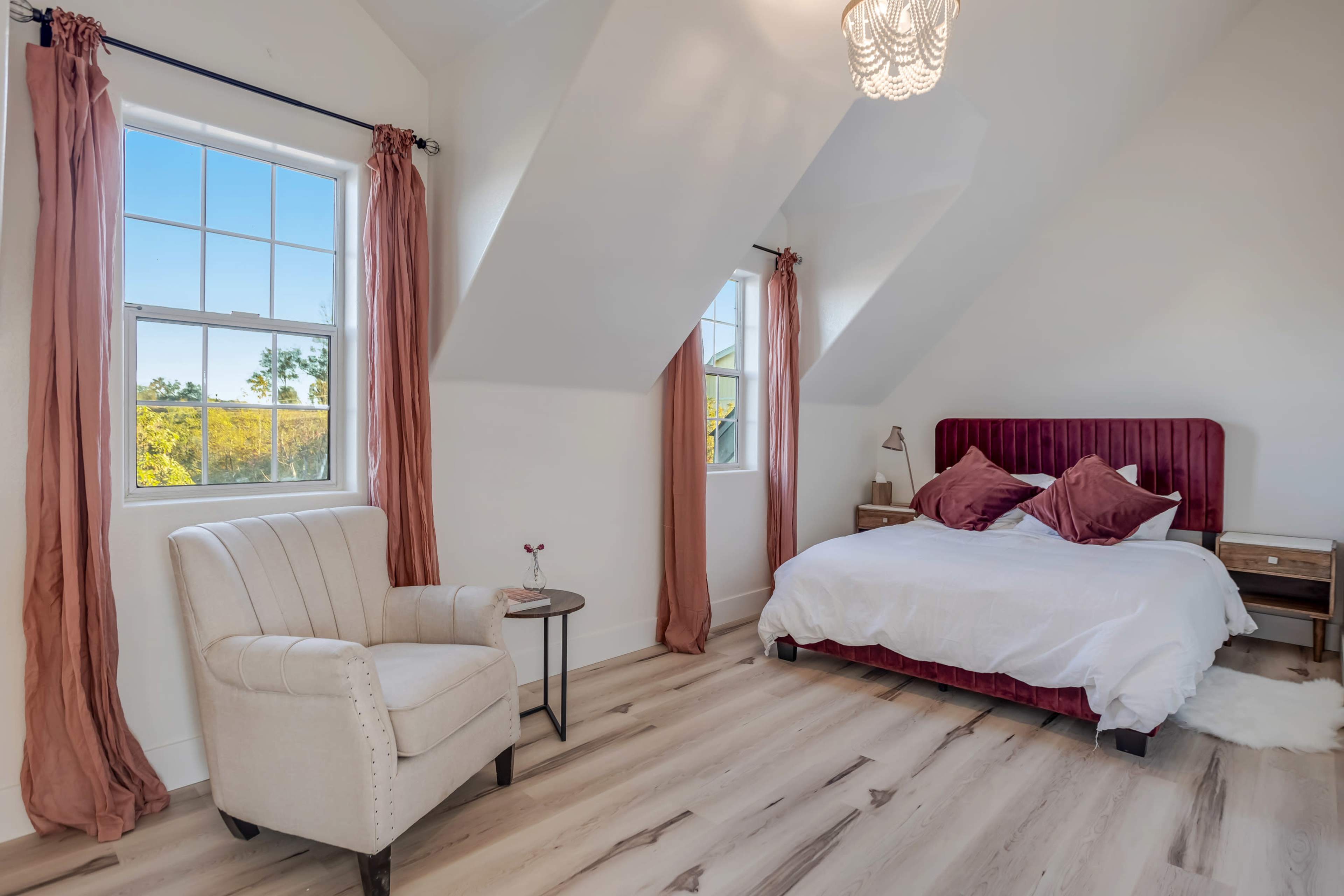 A brightly lit bedroom features a white bed with a burgundy headboard, a small side table, and a cozy armchair near two windows.