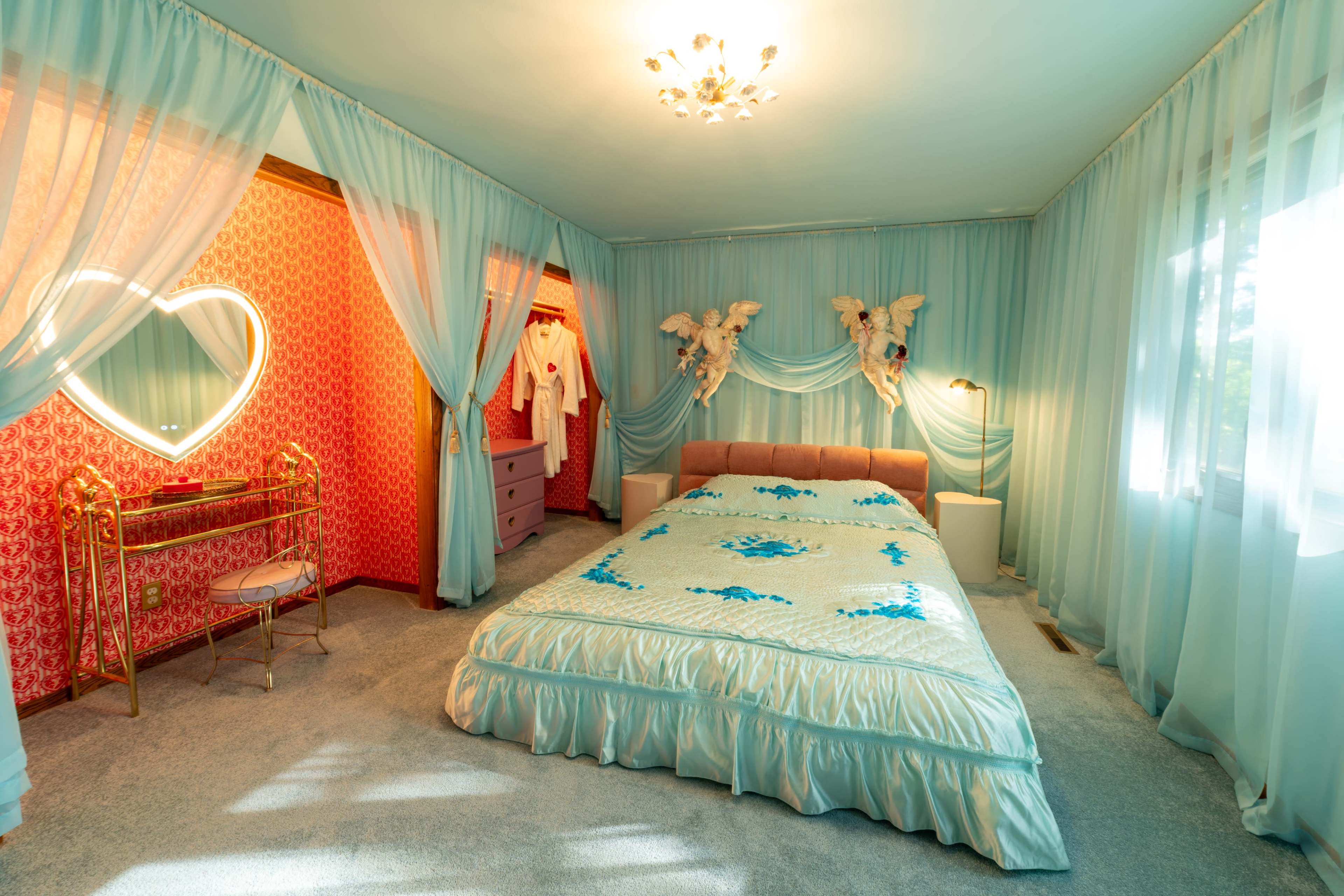 The image shows a bright bedroom featuring a large bed with a blue and white bedspread, surrounded by sheer curtains and adorned with decorative elements, including a heart-shaped mirror and angel figurines.