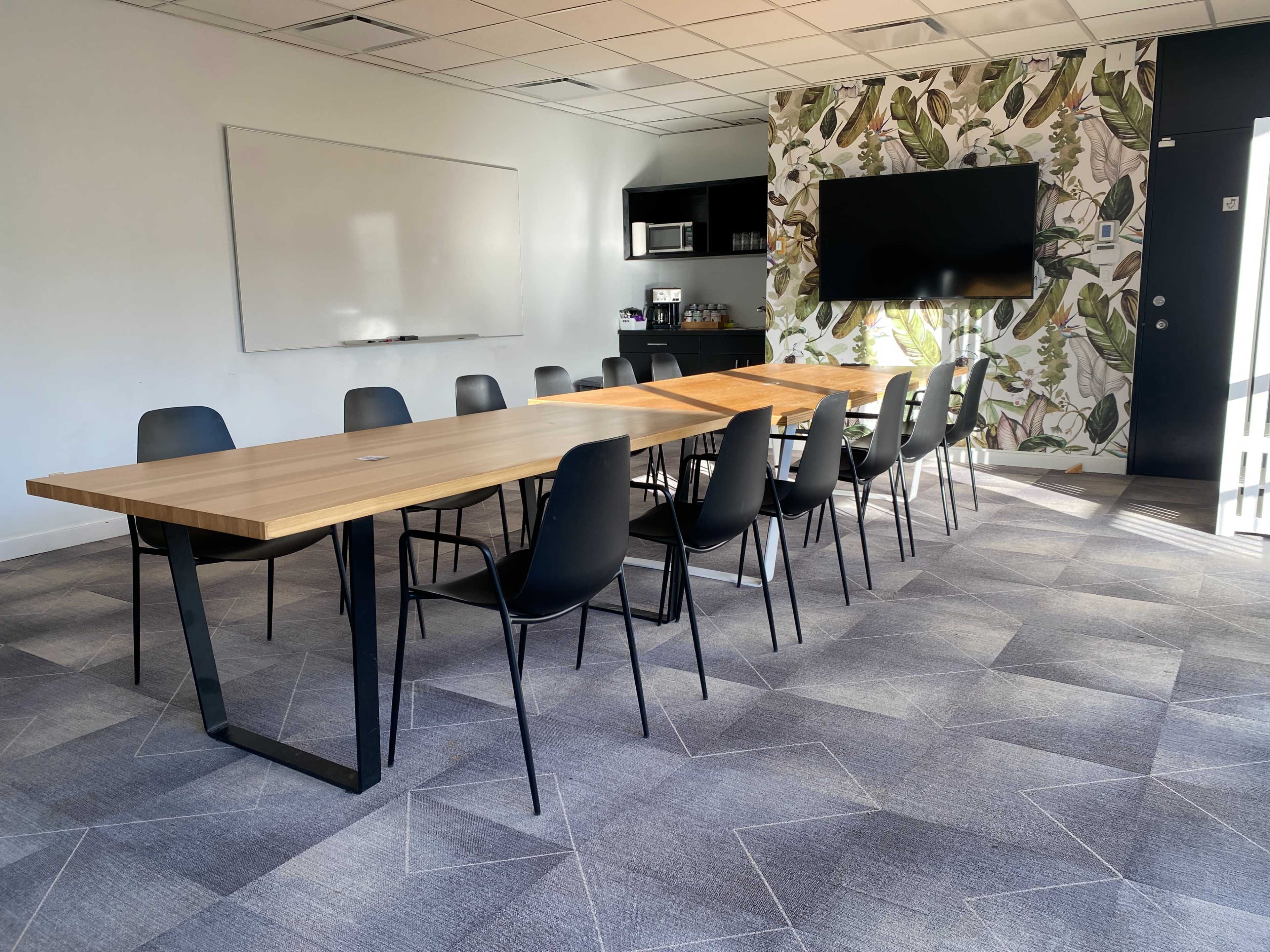 A modern conference room features a long wooden table surrounded by black chairs, with a whiteboard and a large screen on one wall.