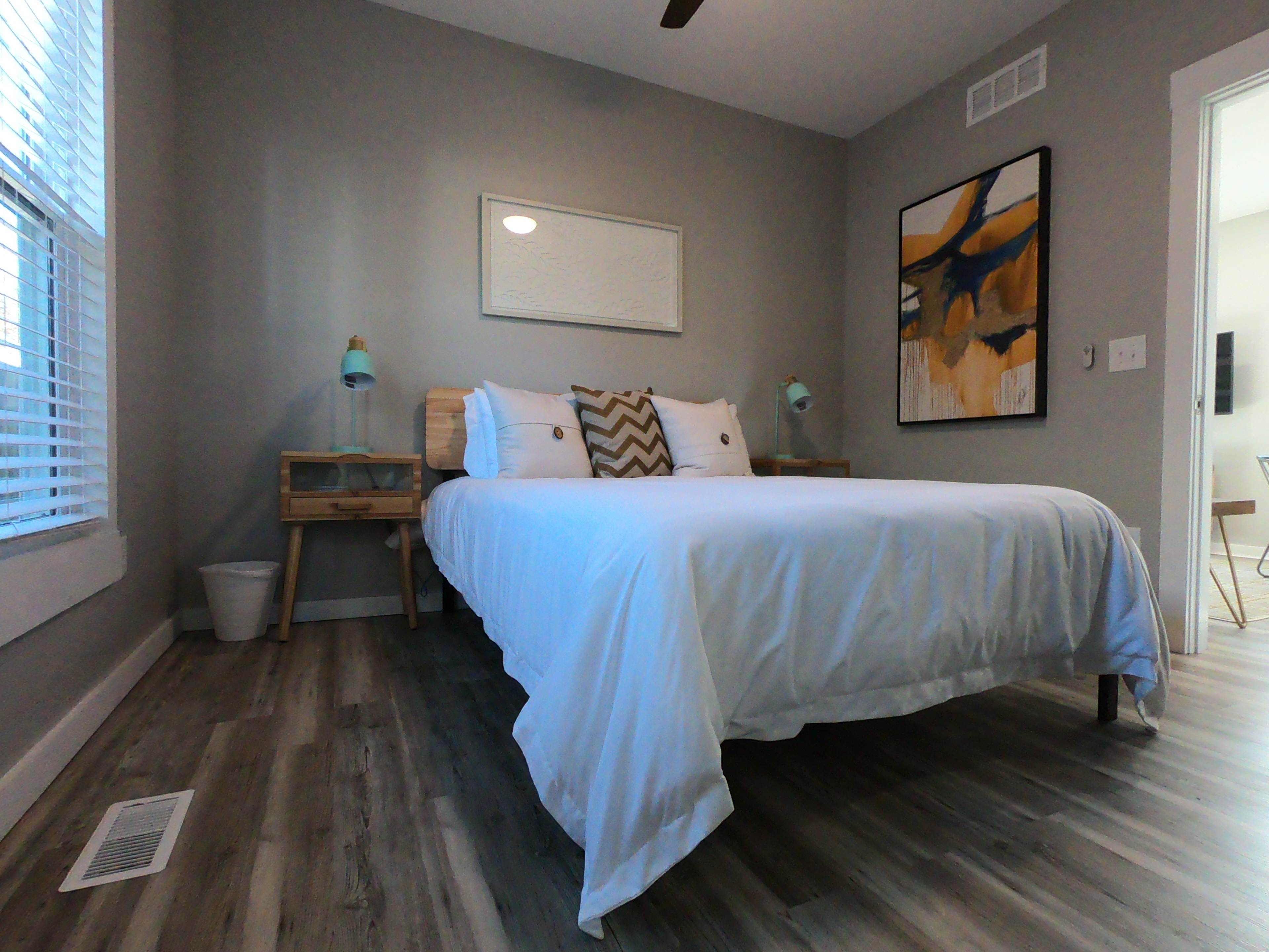 A minimalistic bedroom features a bed with white linens, two bedside tables with lamps, and a large wall art piece.