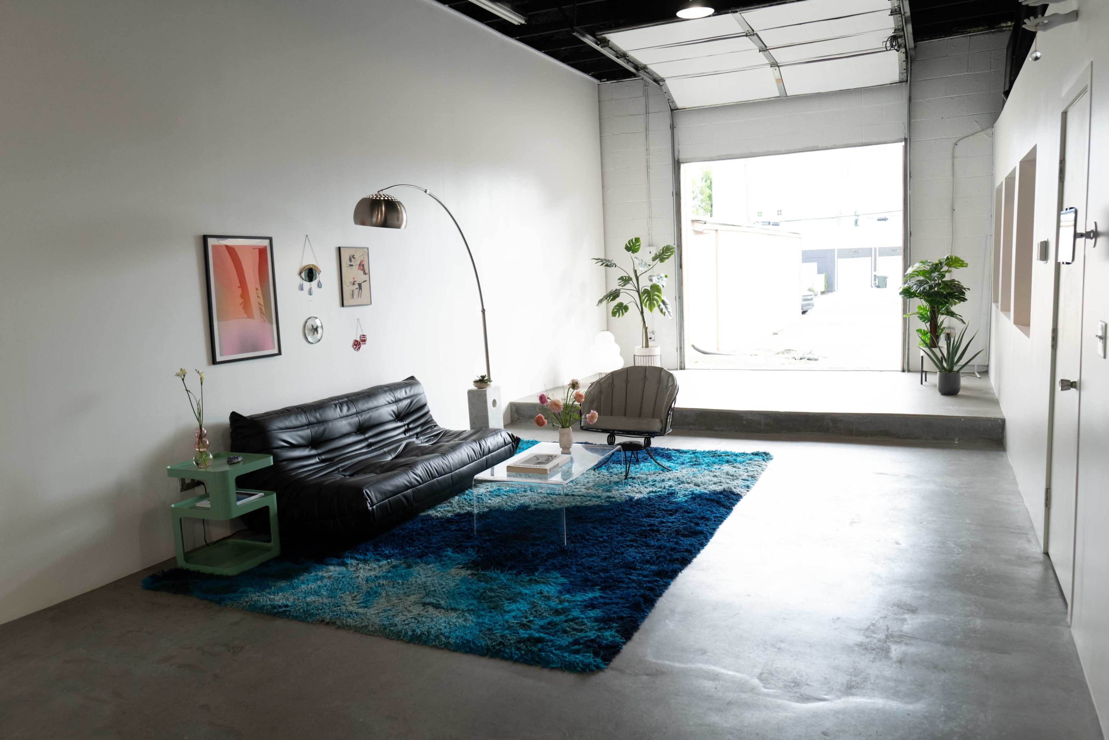 The image features a modern, minimalist living space with a black leather sofa, an arched floor lamp, a glass coffee table, and a blue area rug, situated next to an open garage door letting in natural light.