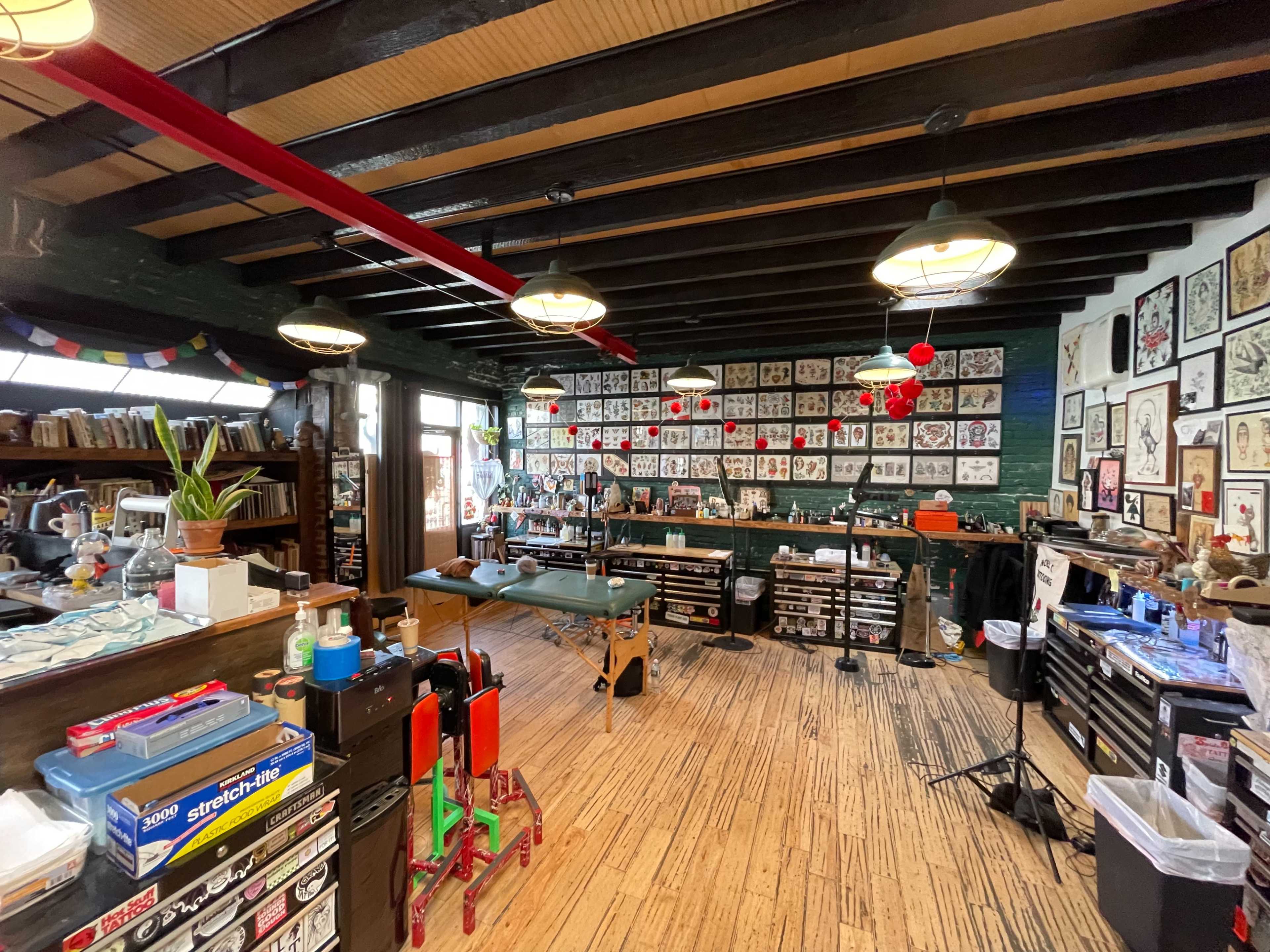 The image shows a well-organized tattoo studio with various artwork and tools displayed on the walls and work surfaces.