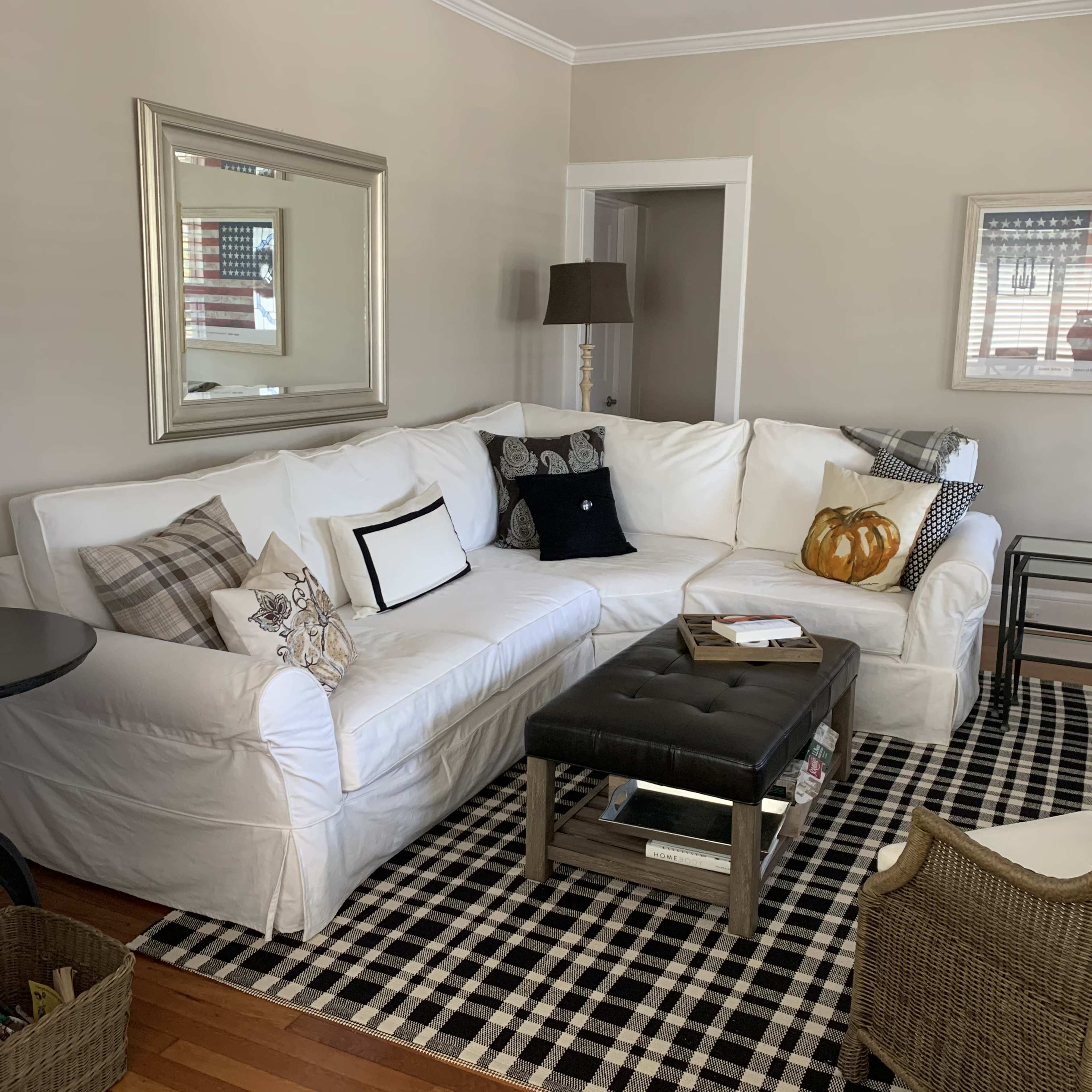 The image shows a cozy living room with a white sectional sofa adorned with various decorative pillows, a black coffee table, and a checkered area rug.