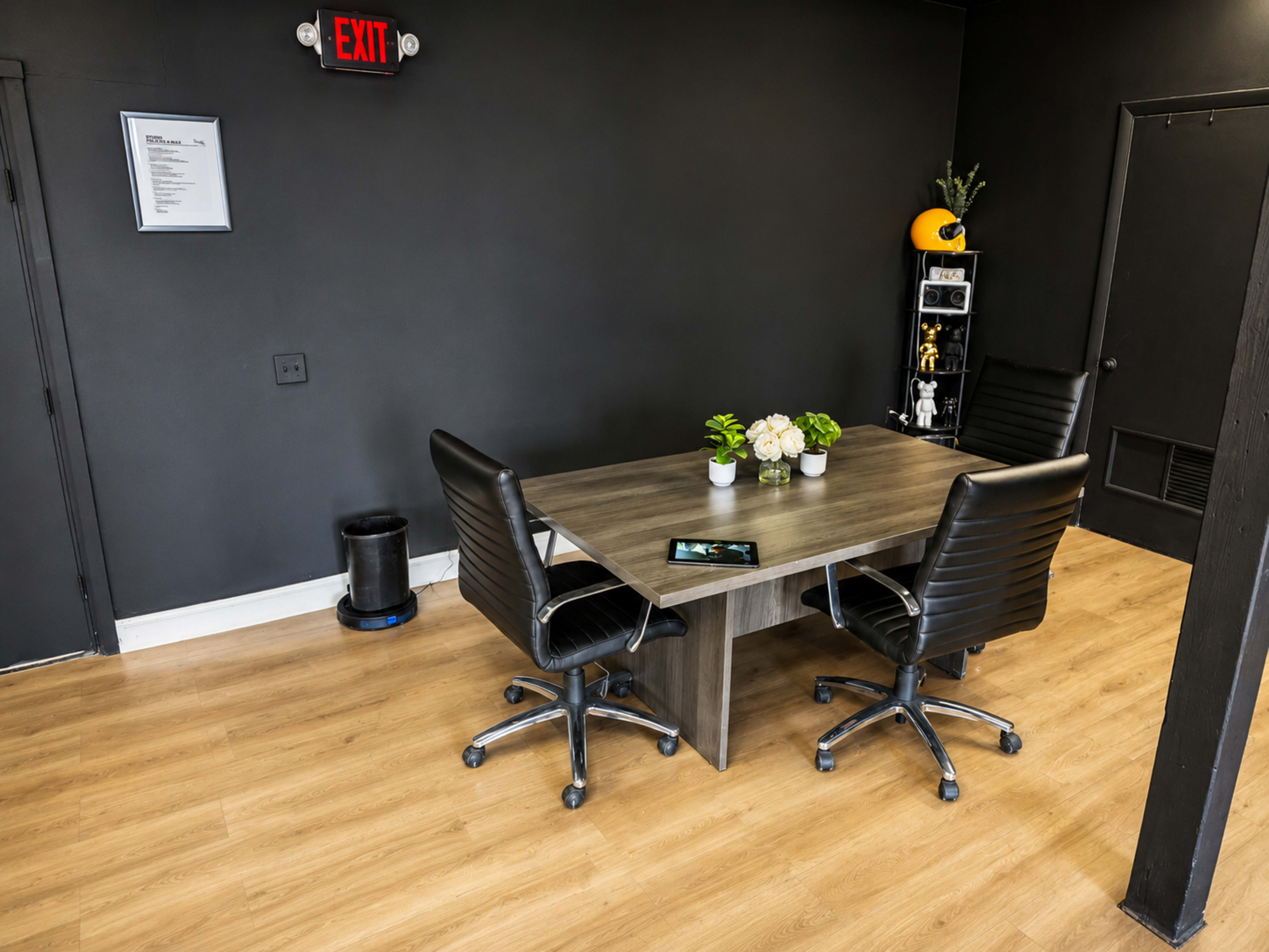 The image shows a small meeting room with a rectangular table, two black office chairs, and a few decorative items on a shelf against a dark-colored wall.