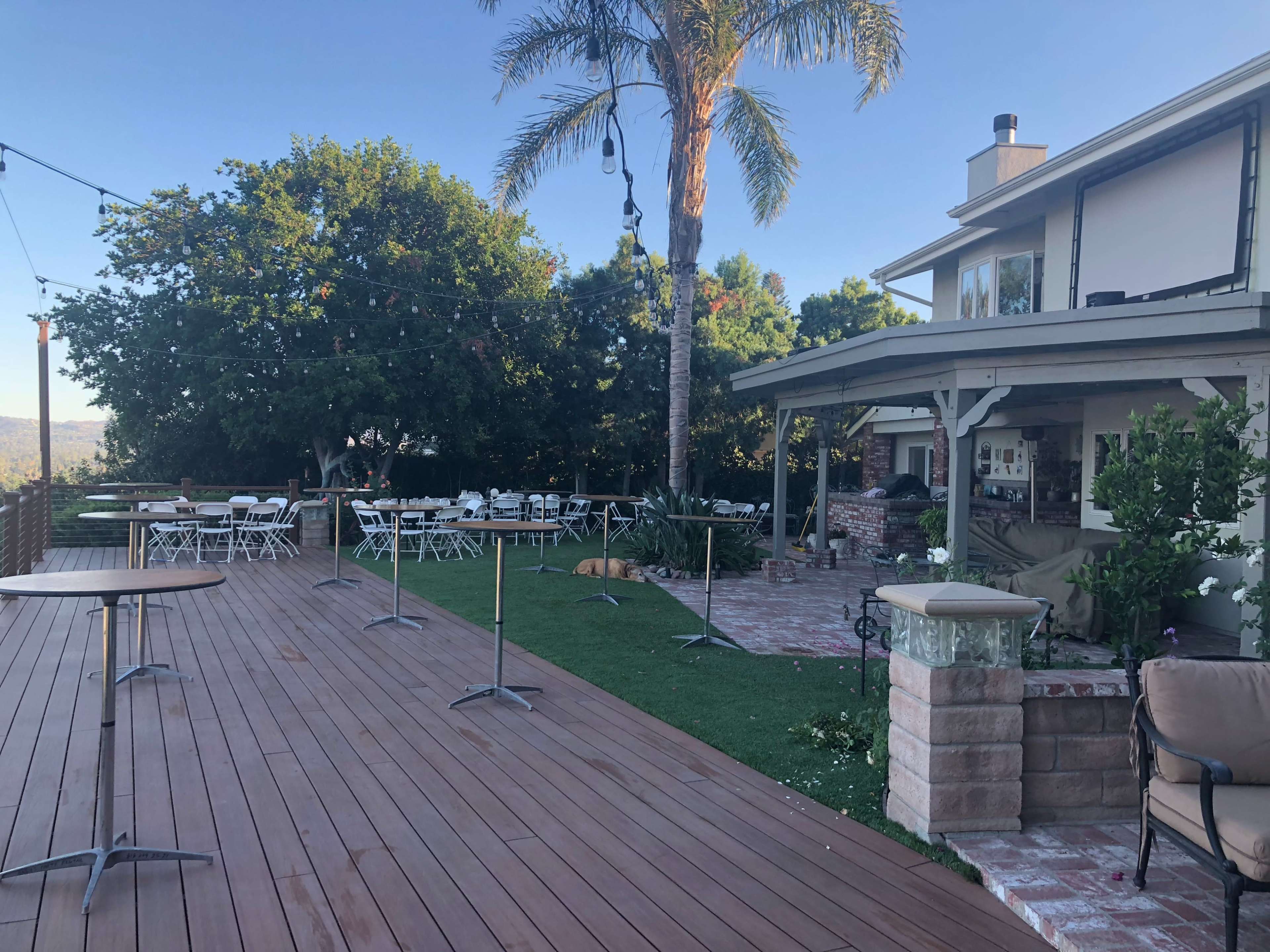 A spacious outdoor deck with folding tables and chairs set up for an event, adjacent to a house and featuring palm trees and greenery.