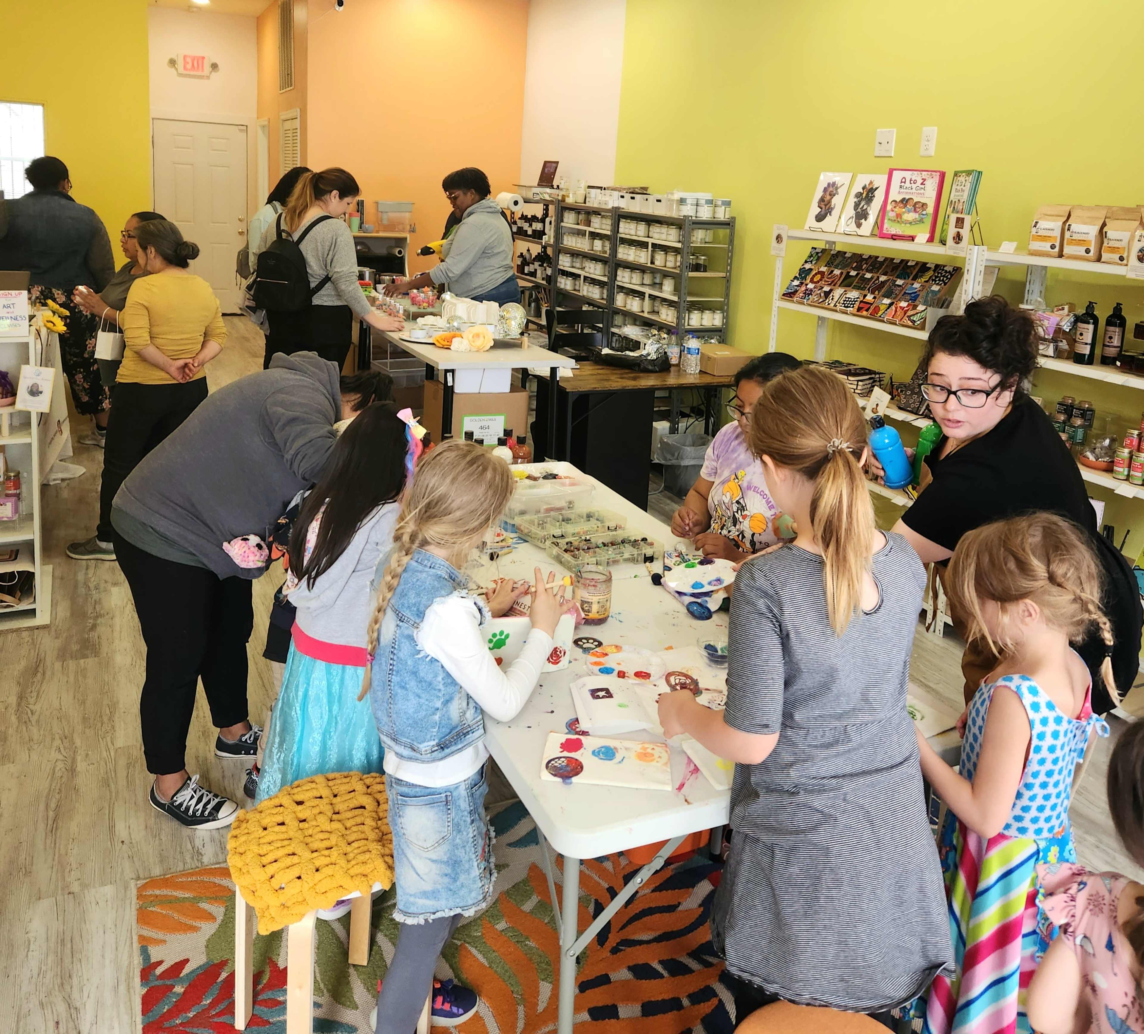 A group of children and adults are engaged in crafting activities at a colorful store, surrounded by shelves filled with various art supplies and products.