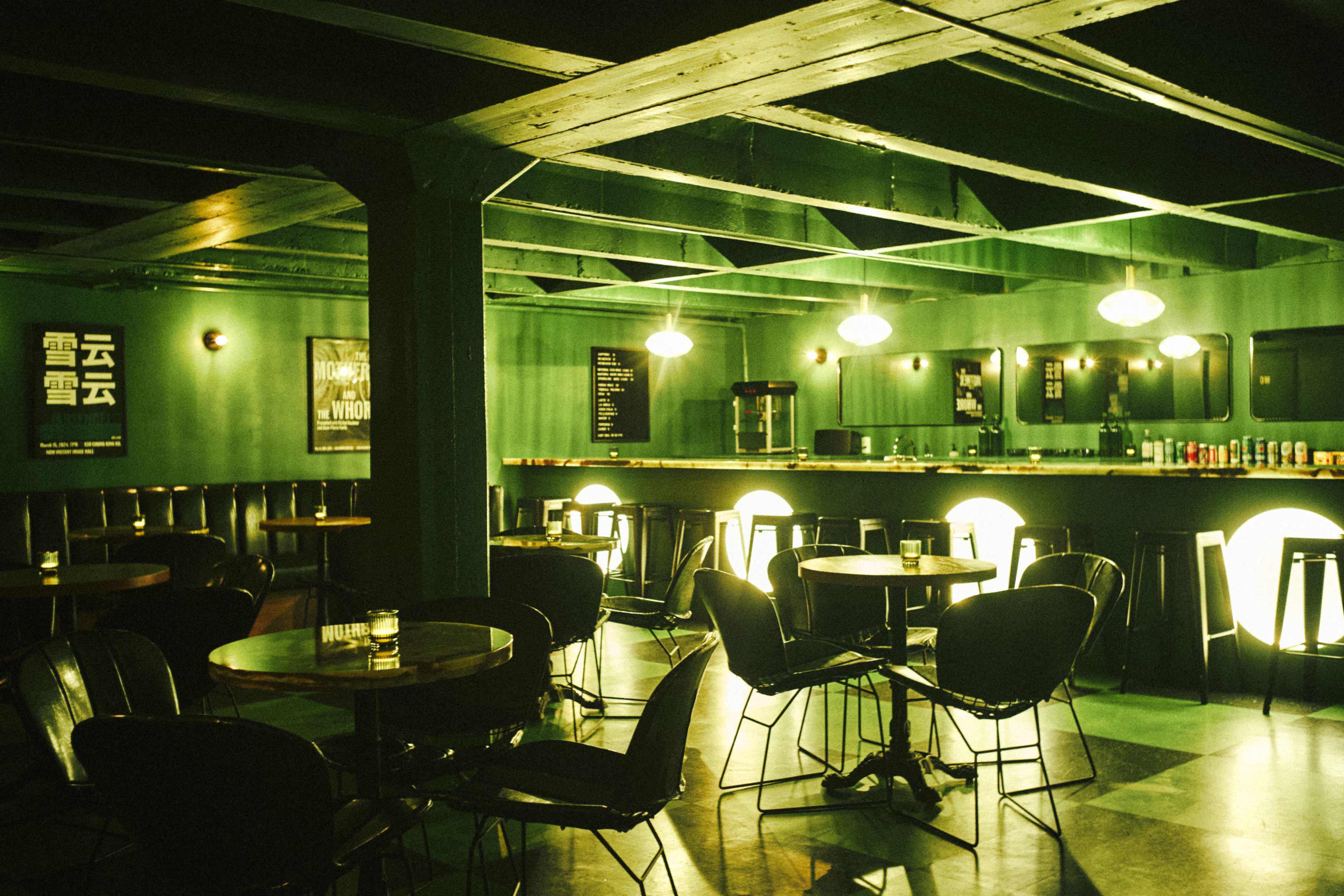 A dimly lit bar features green walls, round tables, and a long counter with illuminated backlighting.