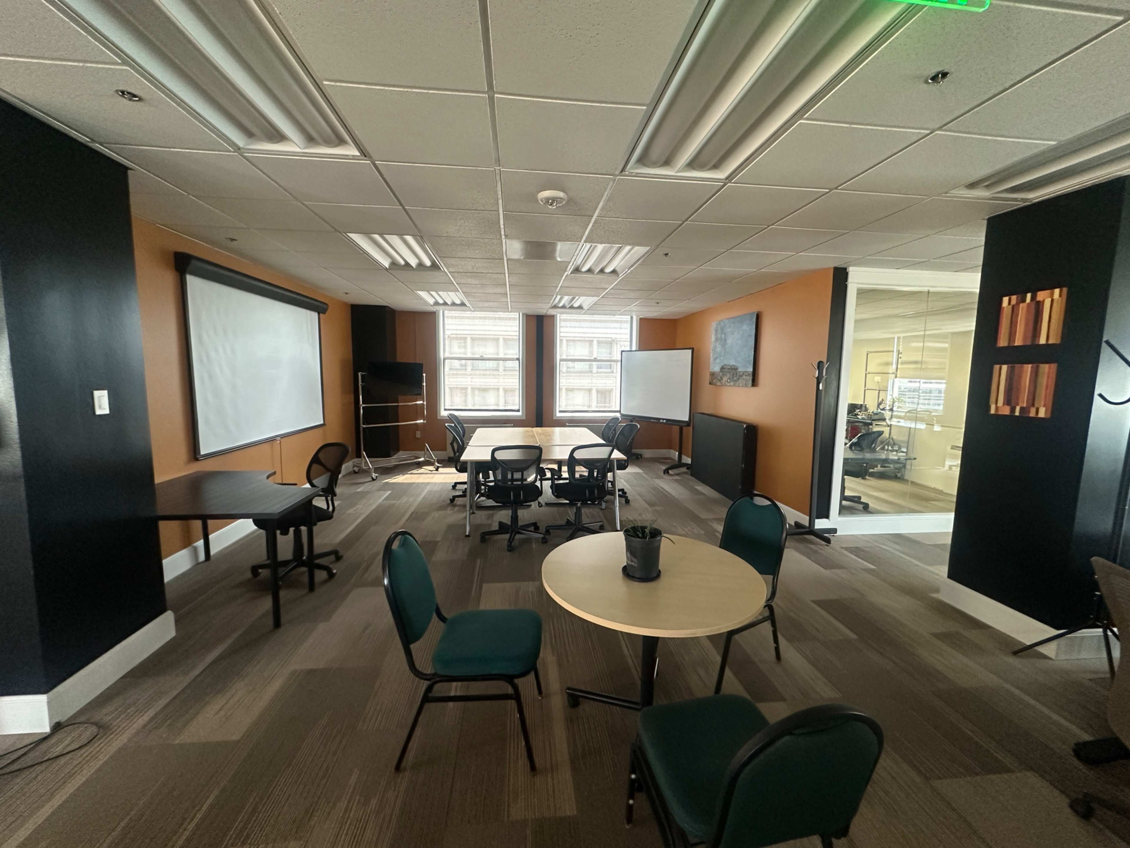 The image shows a conference room with several round tables and chairs arranged around a central area, along with a whiteboard and a projector screen on the wall.