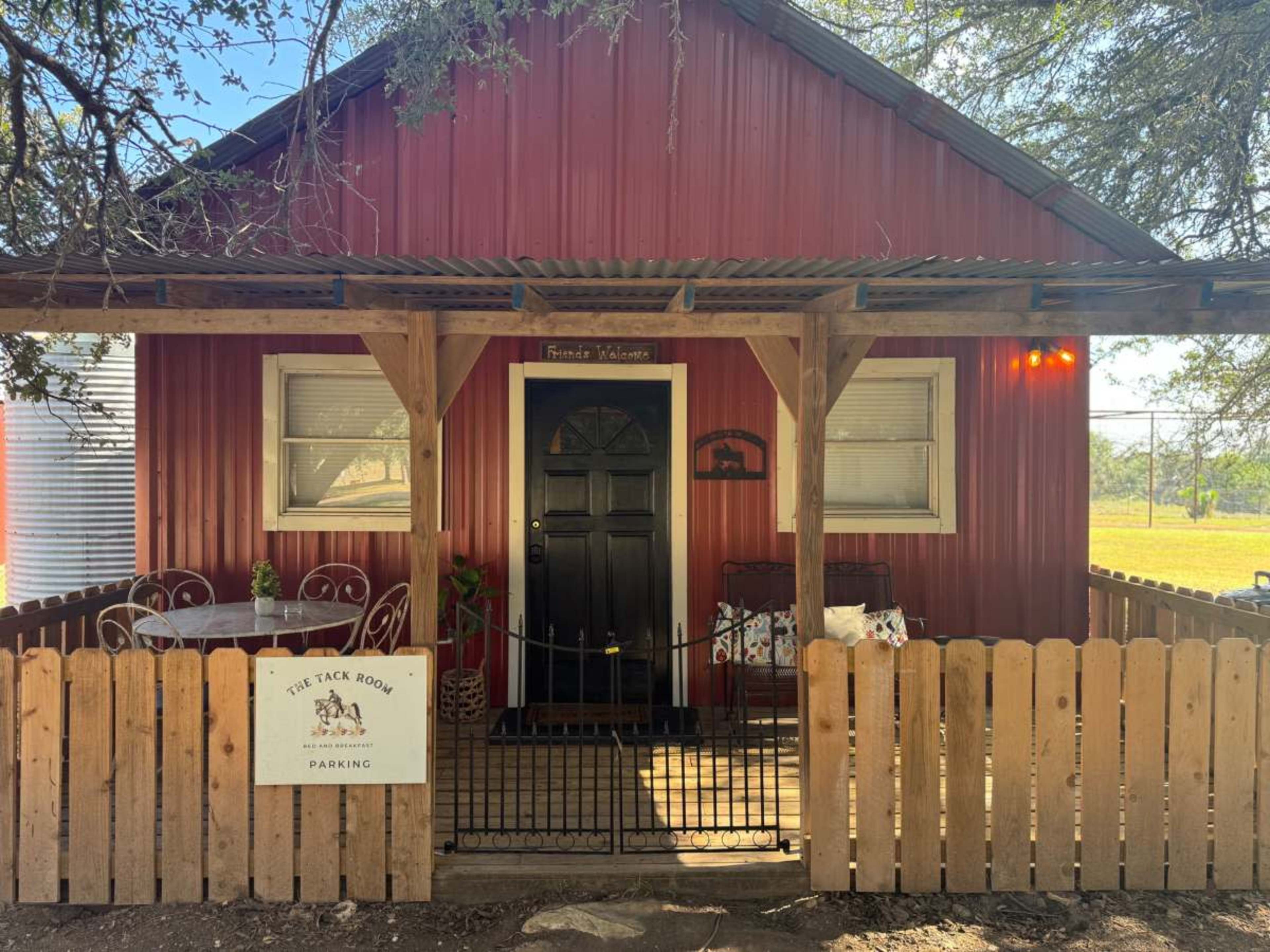 The image shows a small red cabin with a black door, a fenced porch with metal chairs, and a sign reading "The Tack Room - Parking."