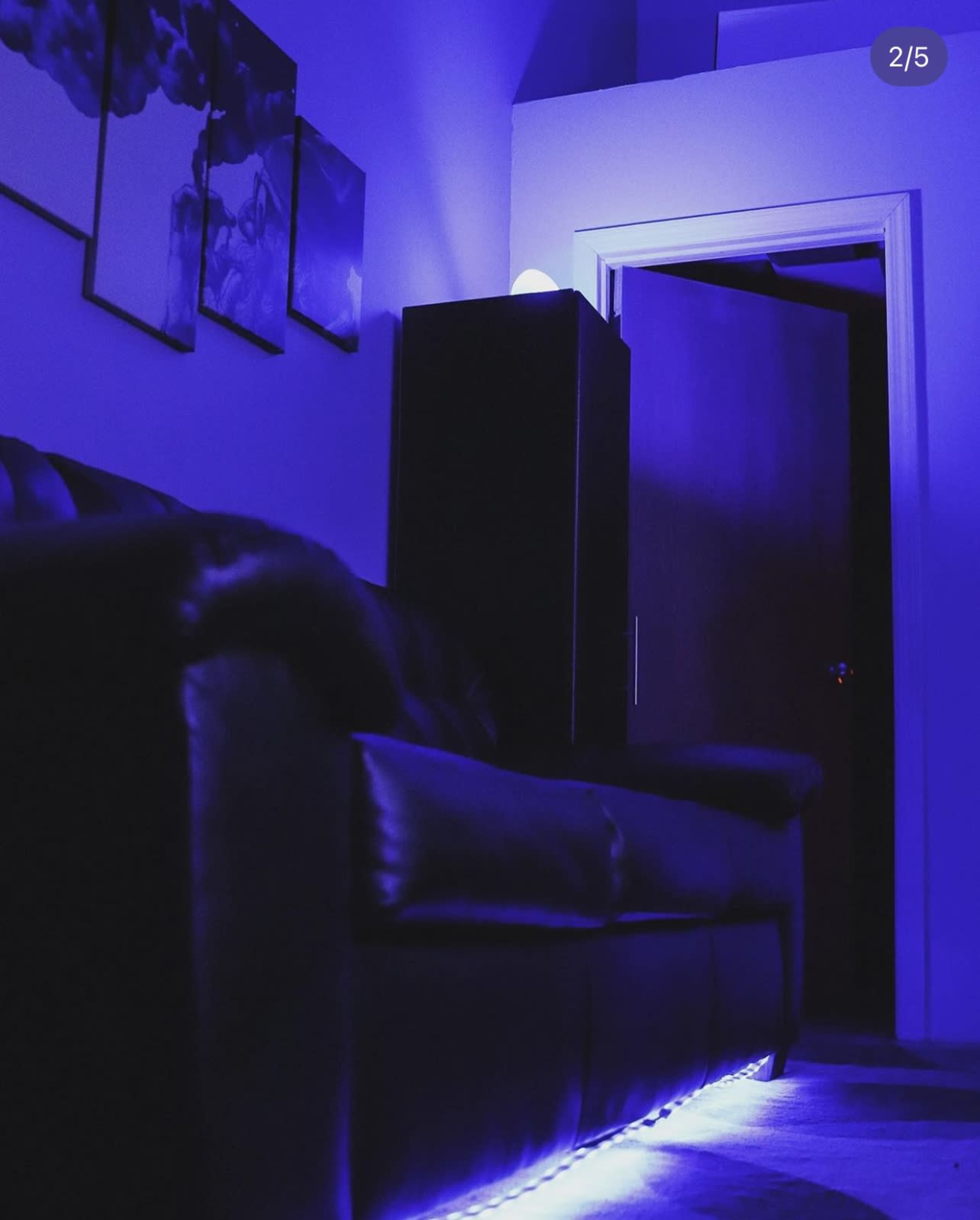 A dimly lit room features a black leather couch with blue lighting underneath, and a door is partially open in the background.