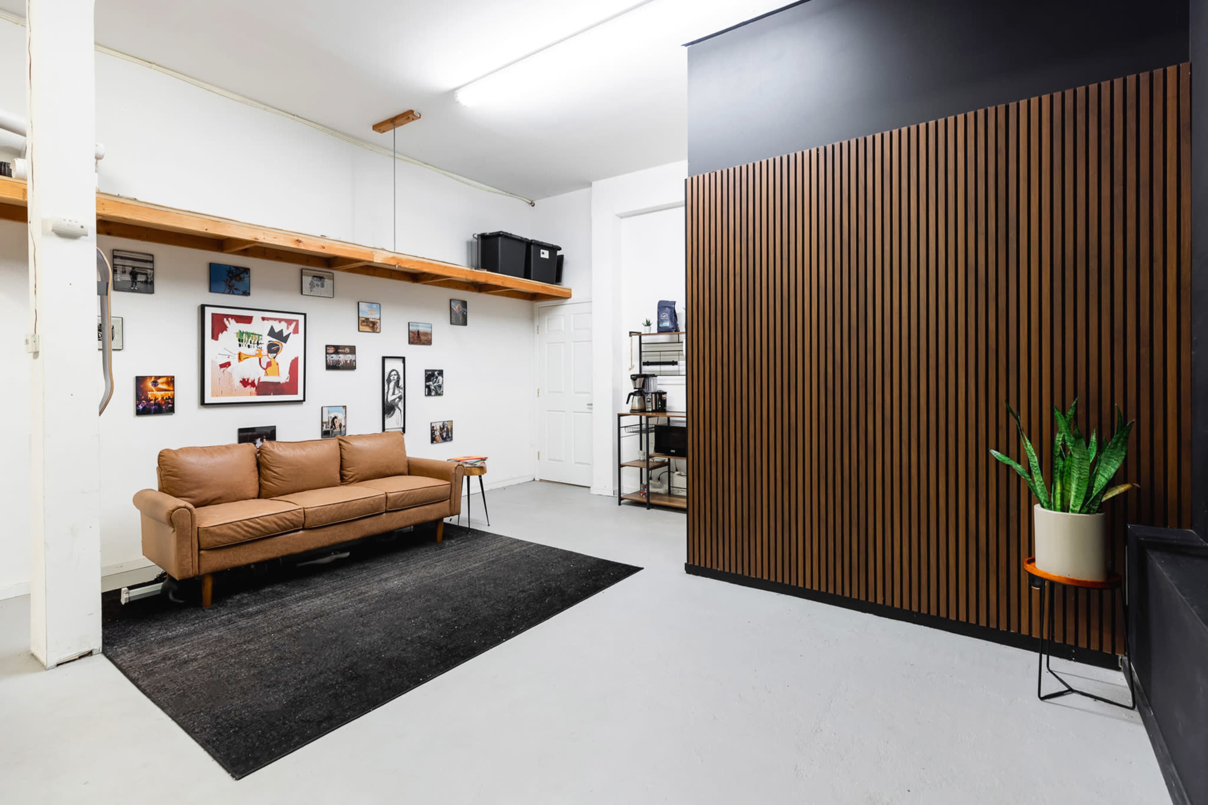 A minimally decorated room with a brown sofa, a black rug, framed pictures on the walls, and a vertical wooden panel next to a potted plant.