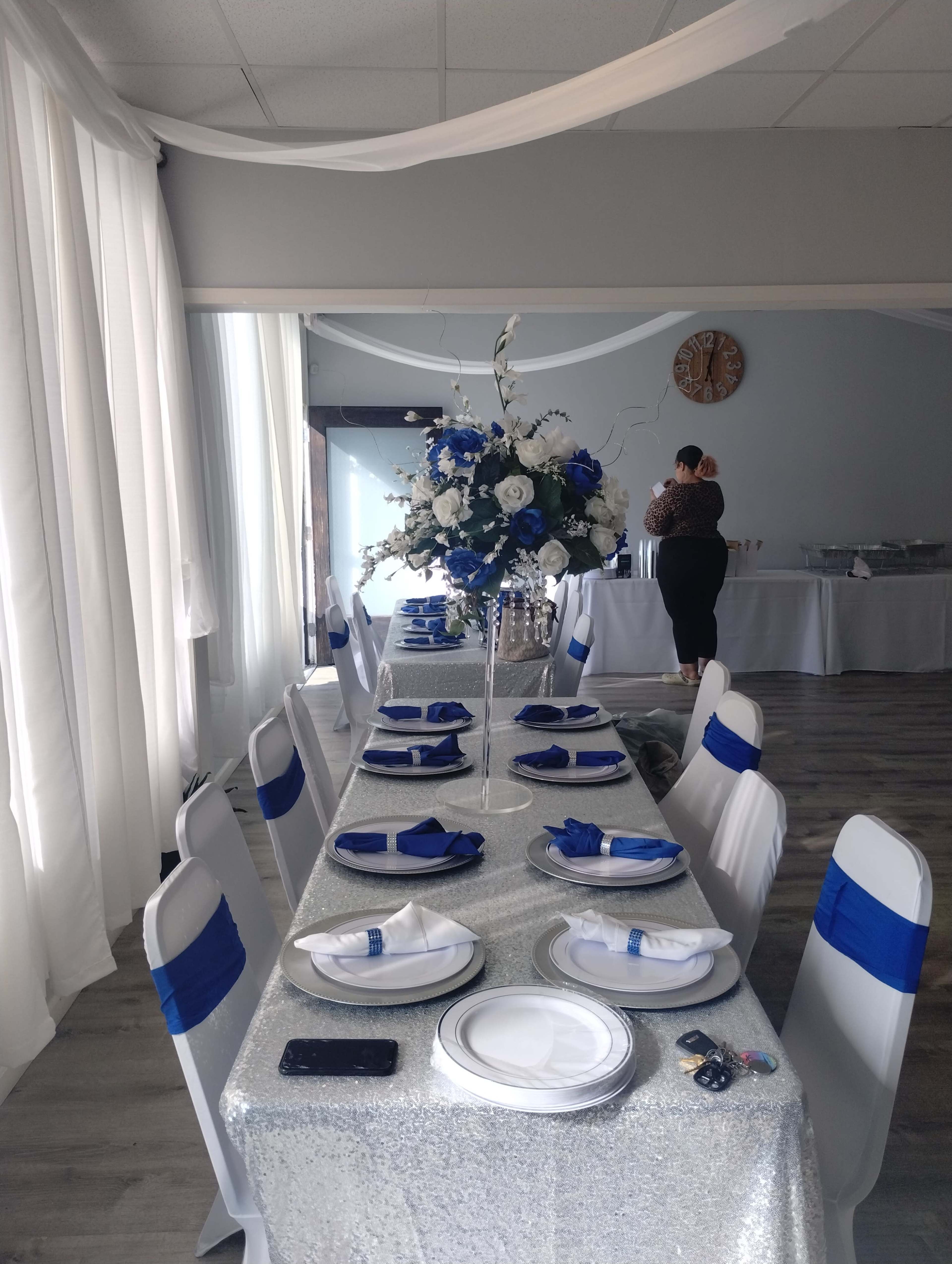 A long table is set with white plates and blue napkins, adorned with a floral centerpiece, in a room decorated with white drapes and a clock on the wall.