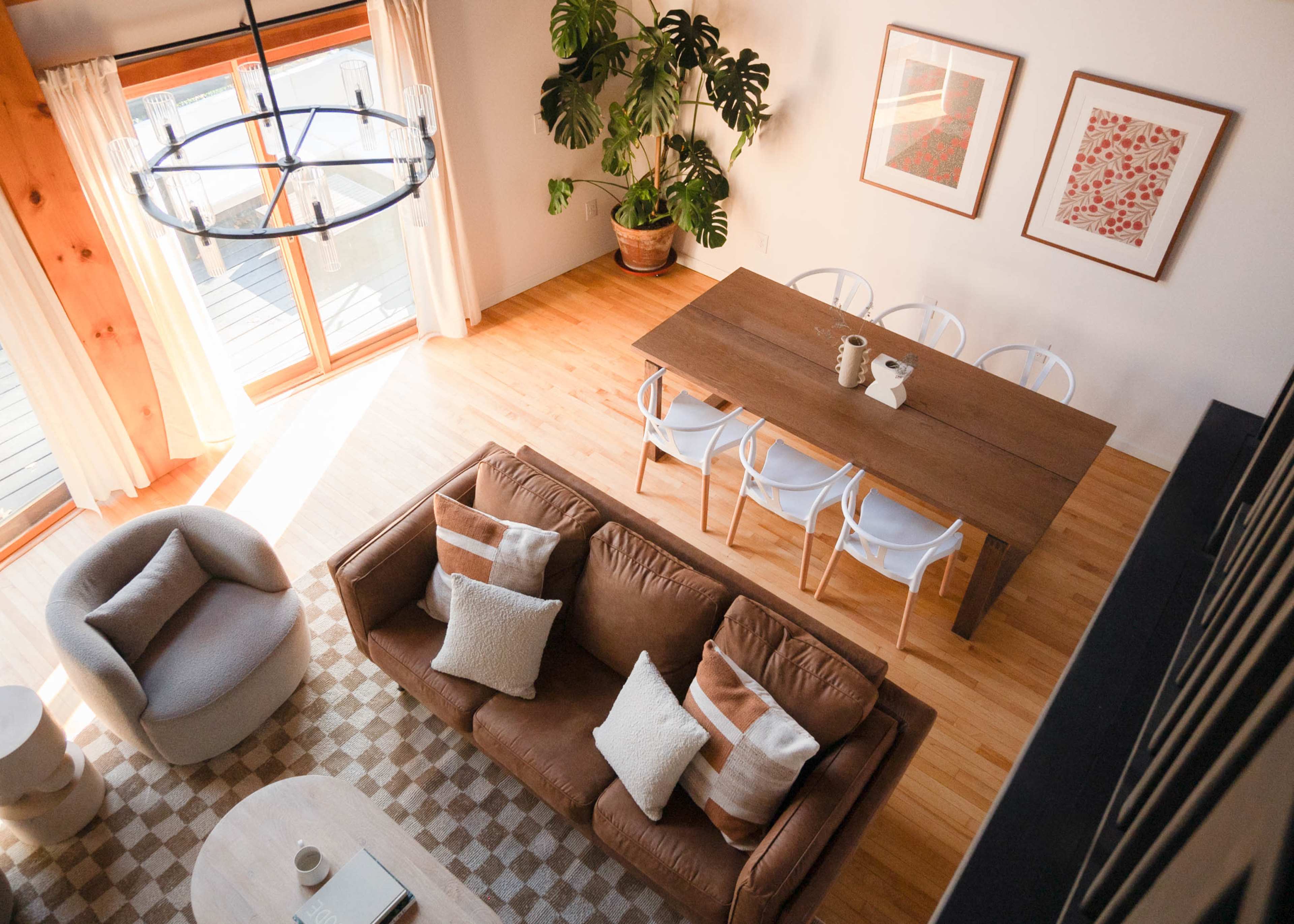 A modern living and dining area with a large wooden table, a brown leather sofa, and potted plants under natural light from large windows.