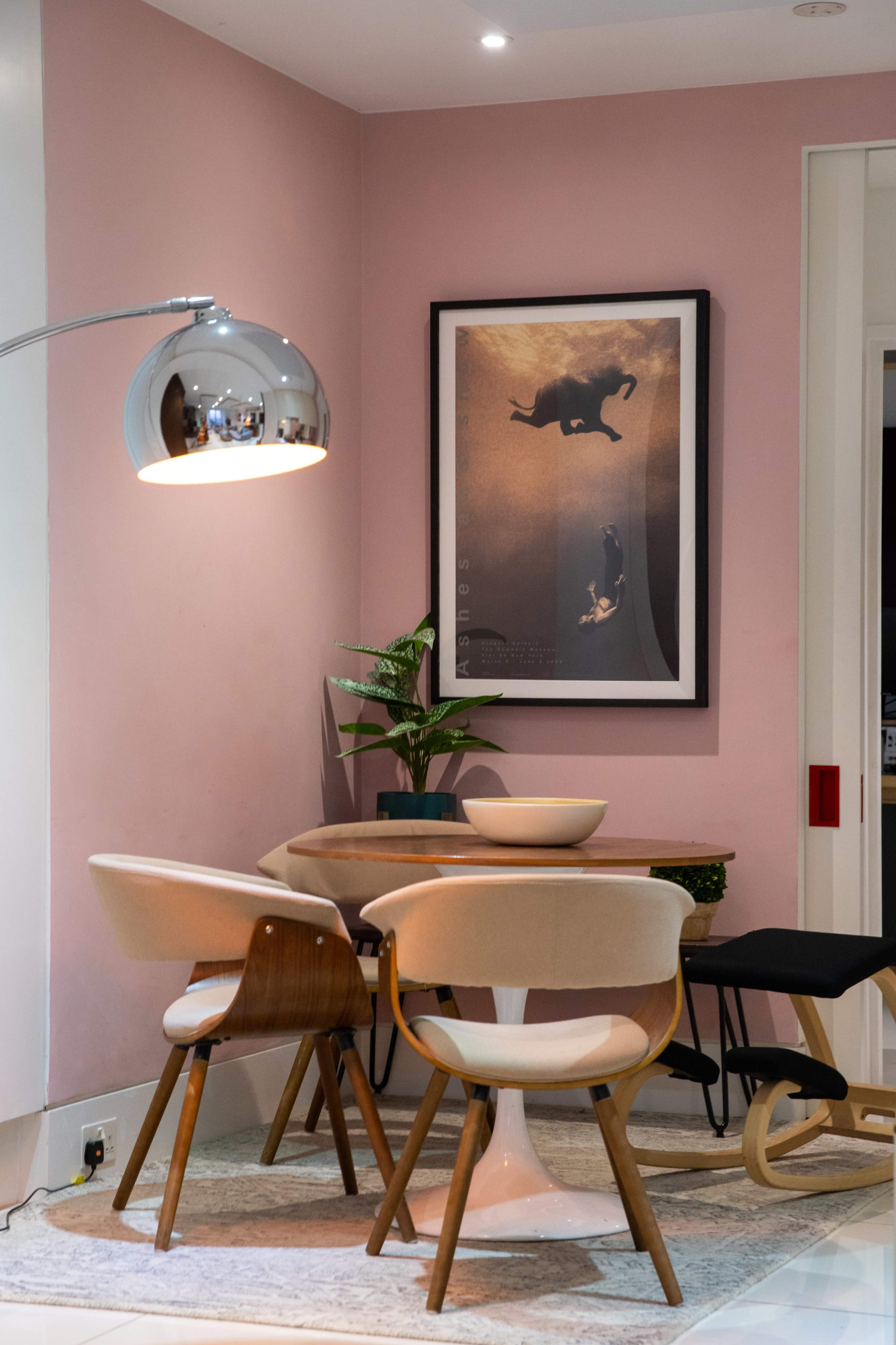 A modern dining area features a round table surrounded by four chairs, a large framed poster on the pink wall, and a floor lamp illuminating the space.