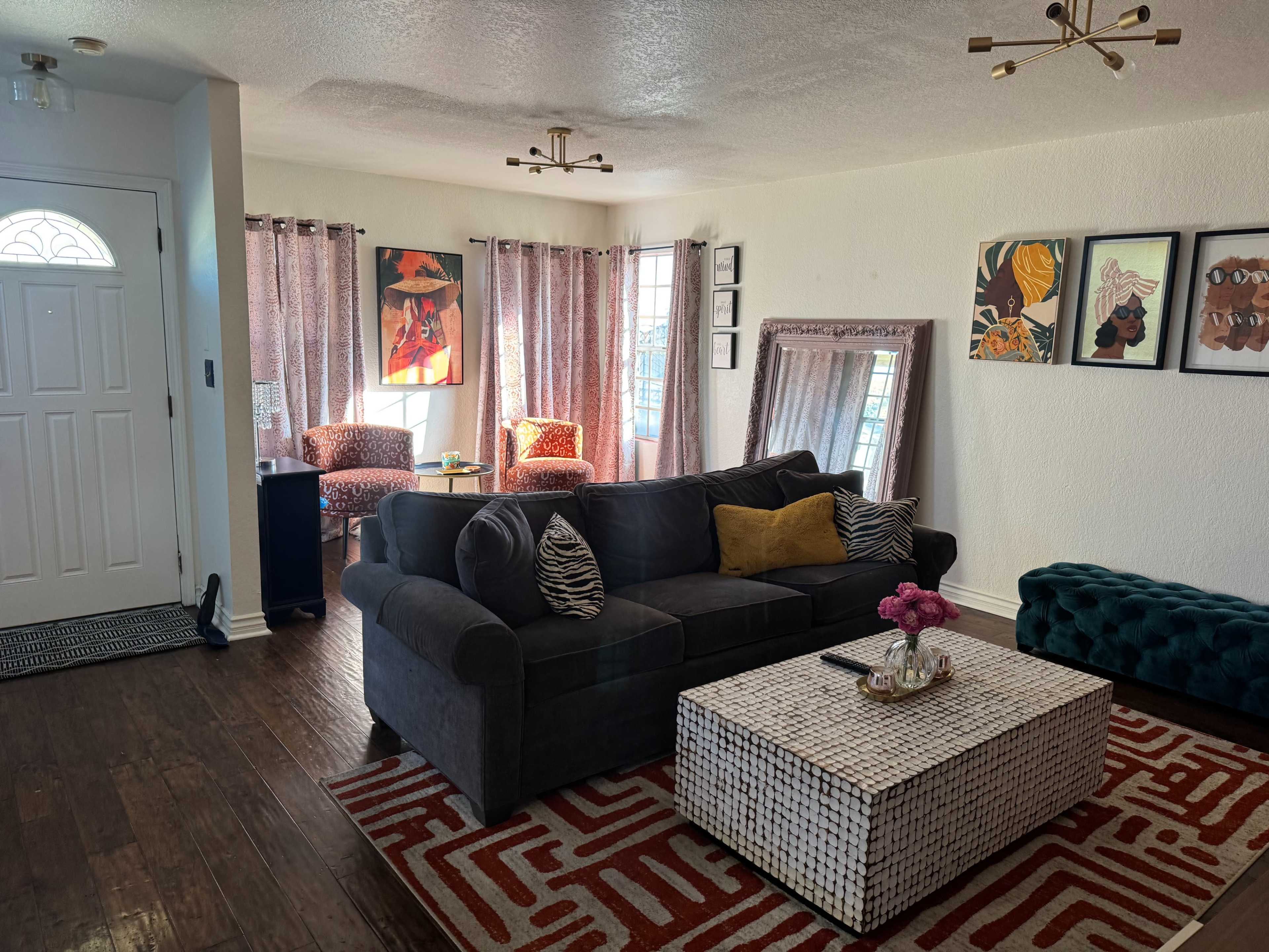 The image shows a living room with a dark gray sectional sofa, a patterned area rug, a coffee table, and large windows dressed with curtains, offering a cozy seating arrangement.