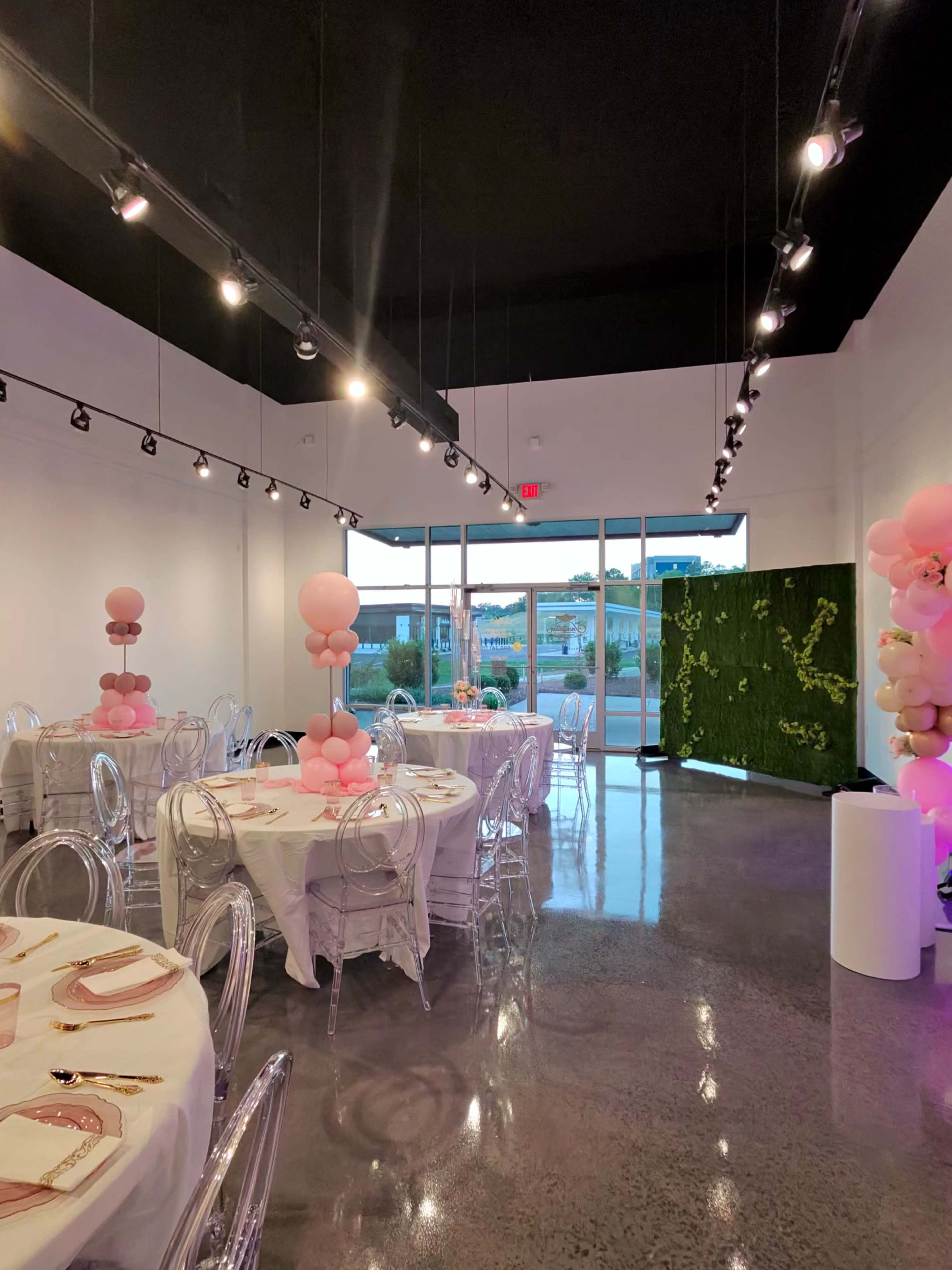 The image shows a modern event space with round tables set for dinner, featuring pink balloon decorations and a green wall in the background.