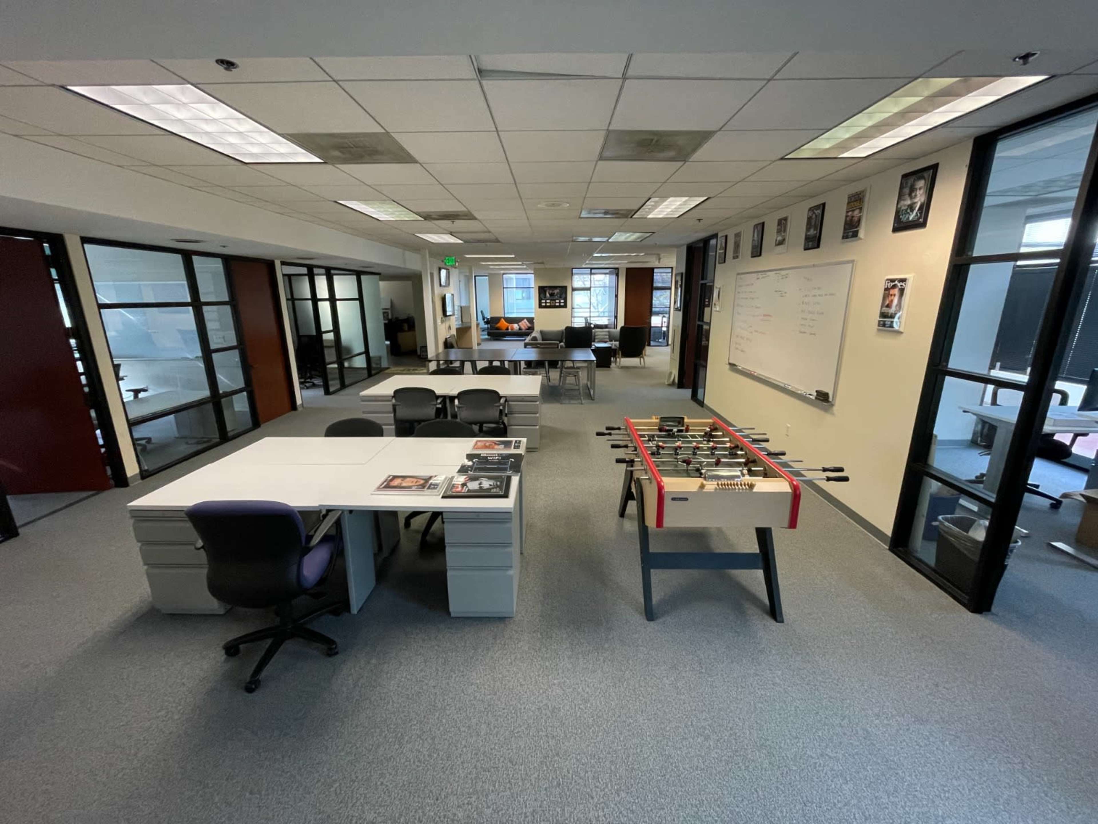 An office space with rows of desks, a foosball table in the center, and glass-walled meeting rooms in the background.