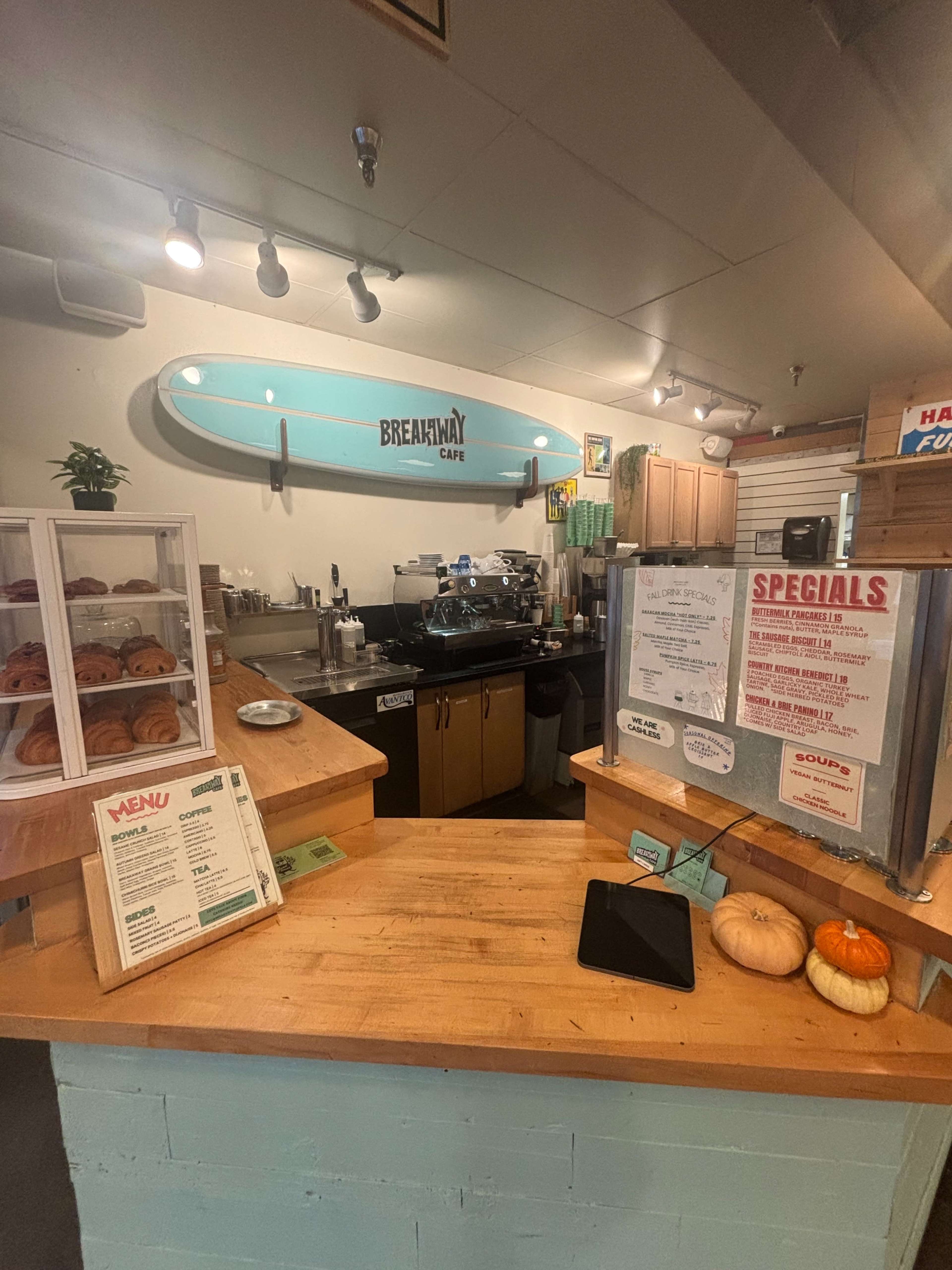 The image shows the interior of a café featuring a surfboard on the wall, a coffee machine on the counter, and a display of baked goods.
