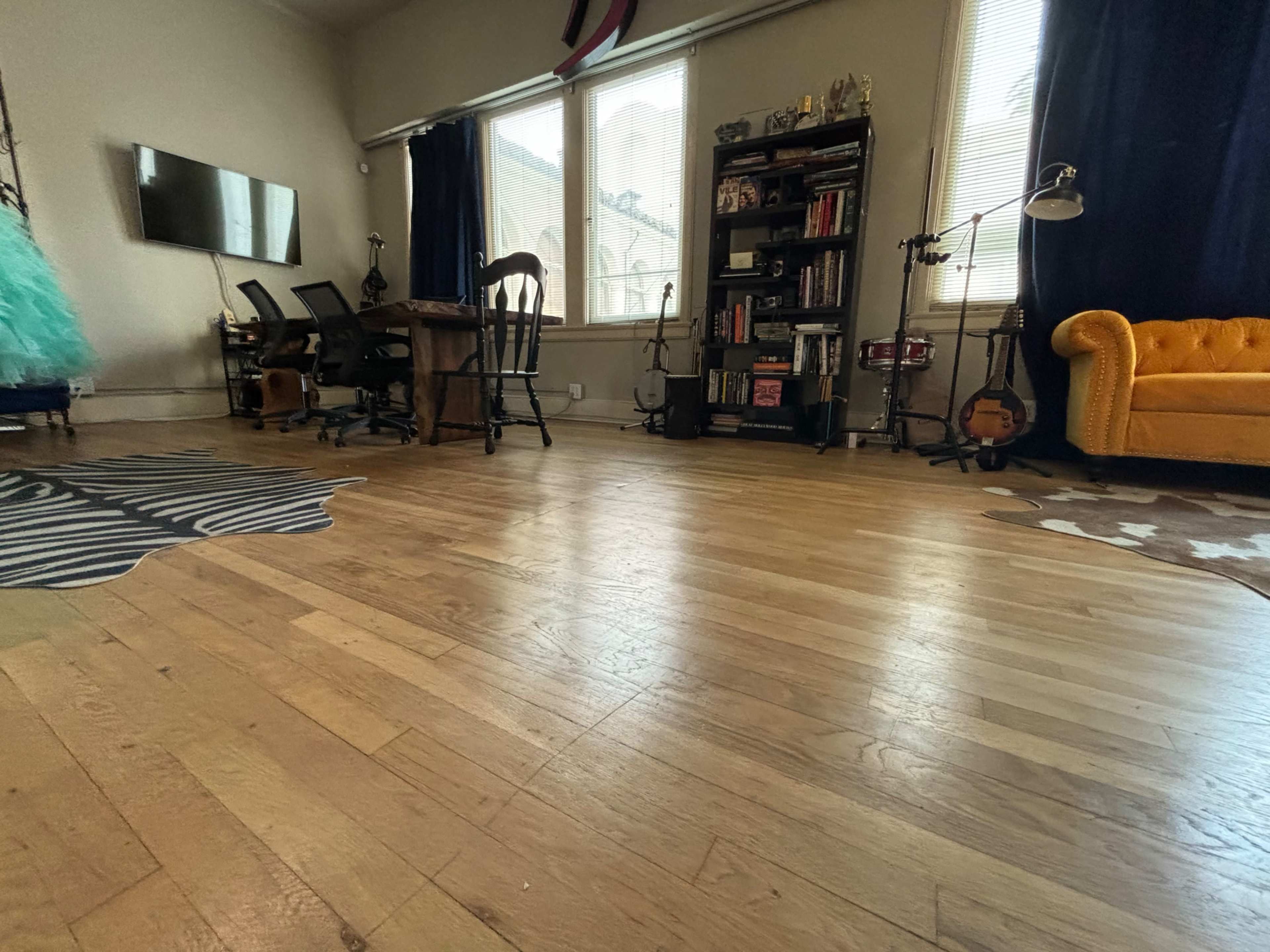 The image shows a room with wooden flooring, a yellow couch, a bookshelf, a desk with chairs, and various musical instruments.