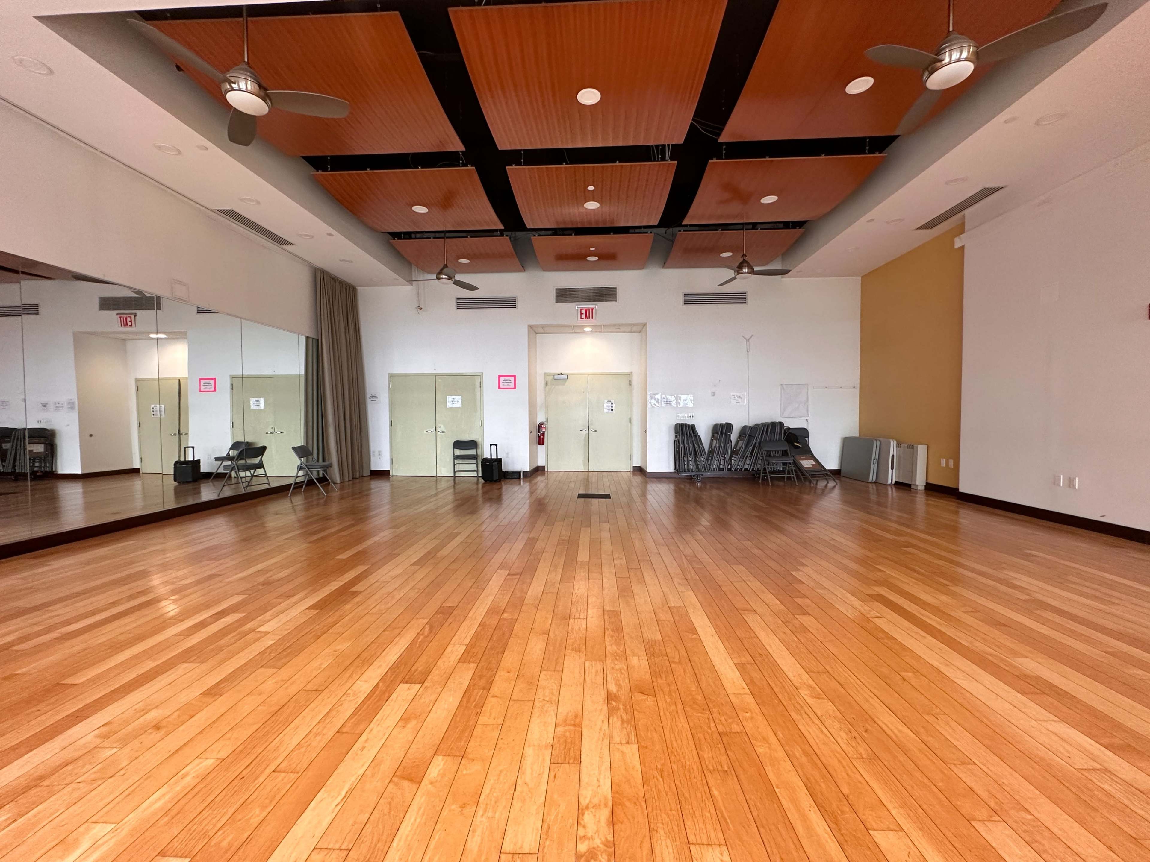 A spacious wooden-floored room with mirrors, ceiling fans, and a few folding chairs along the walls.