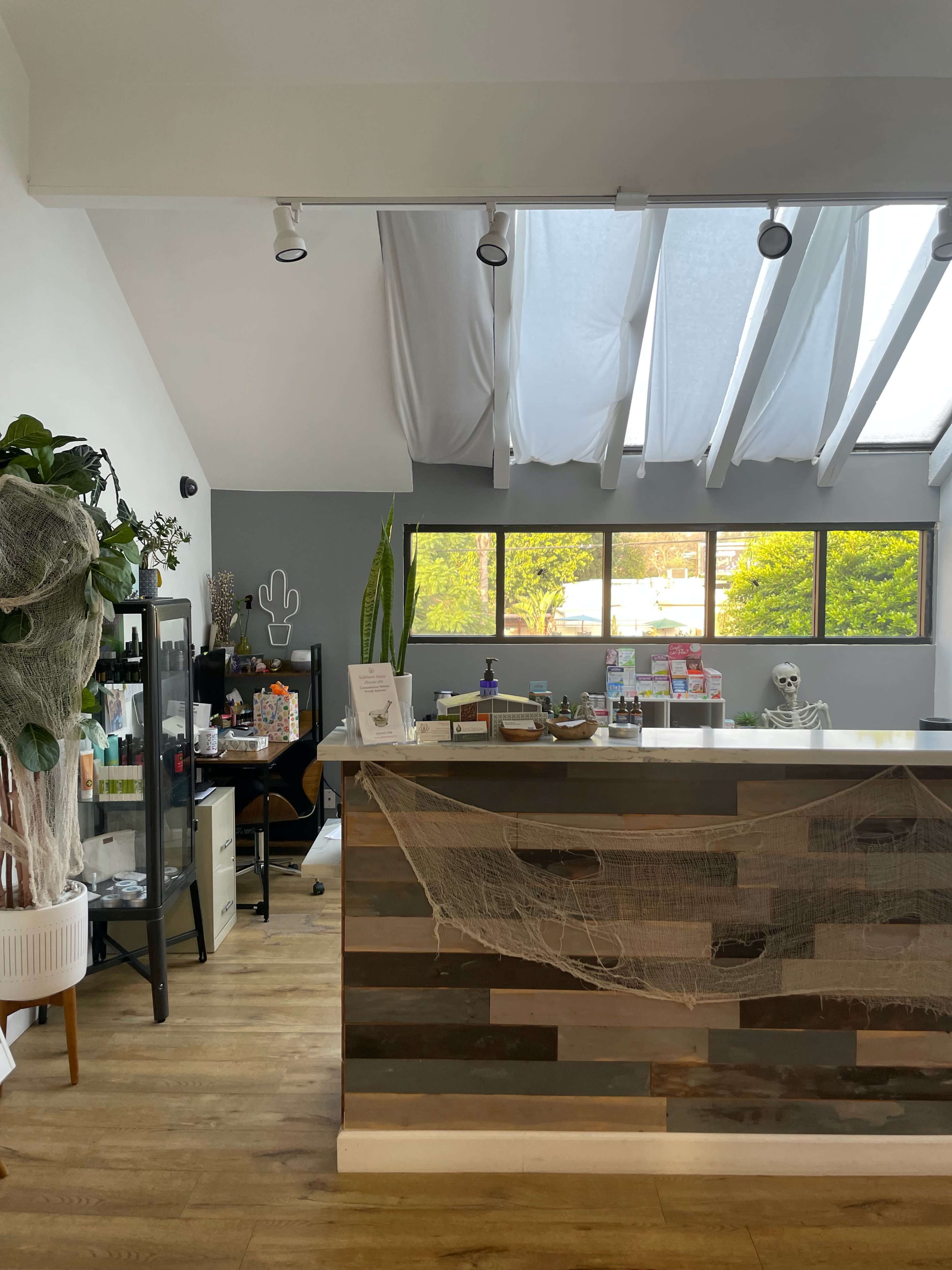 A modern reception area with a wooden front desk, a skeleton decoration, plants, and large windows allowing natural light to enter.