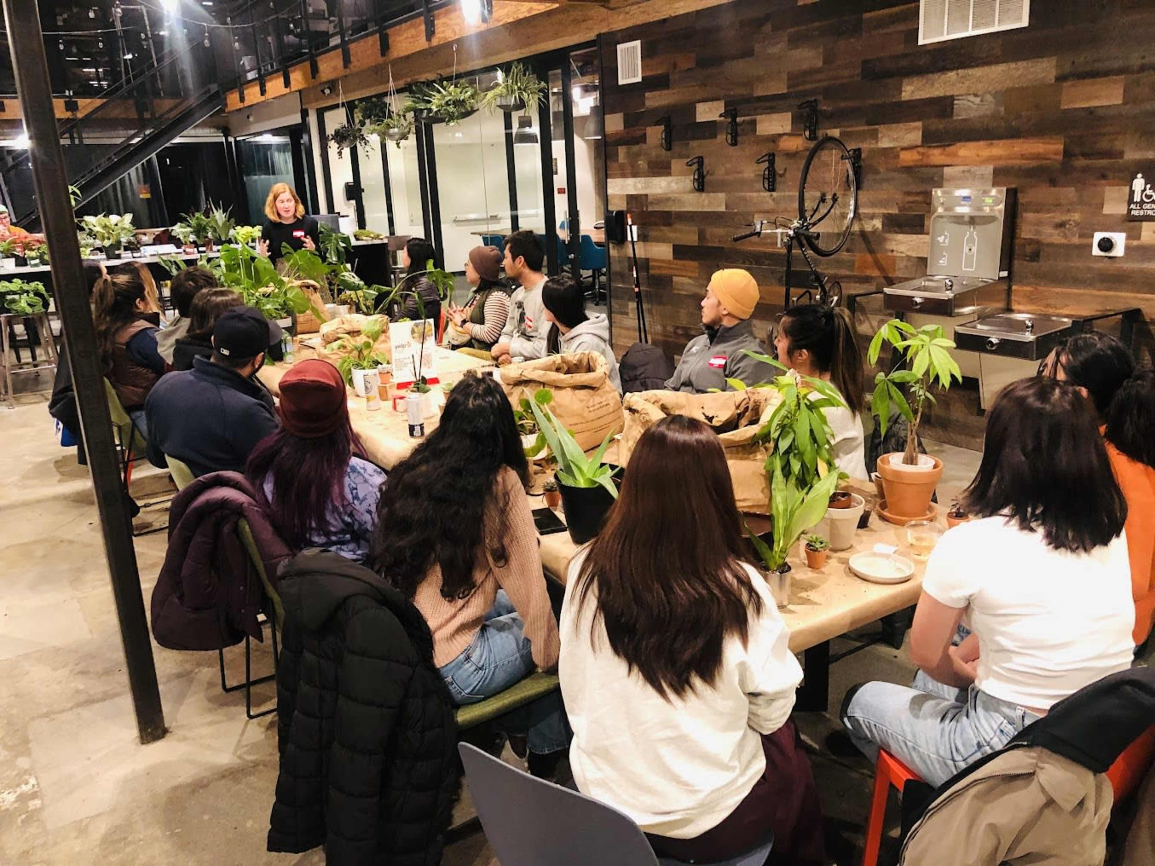 A group of people sits around a long table in a plant-filled space, listening to a speaker at the front.