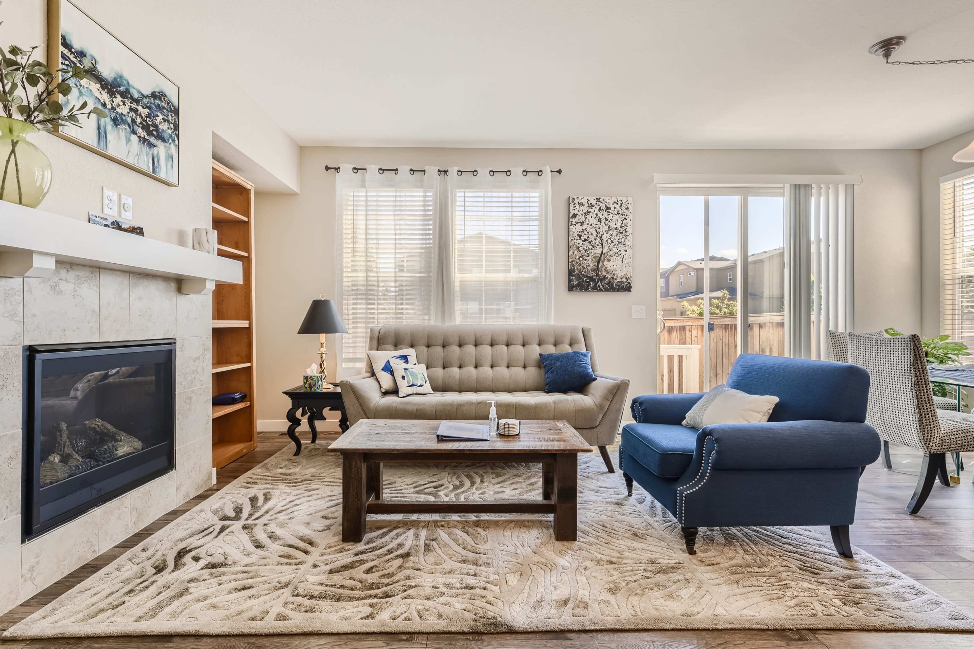 A living room with a beige sofa, a blue armchair, a coffee table on a patterned rug, and a fireplace, illuminated by natural light from large windows.