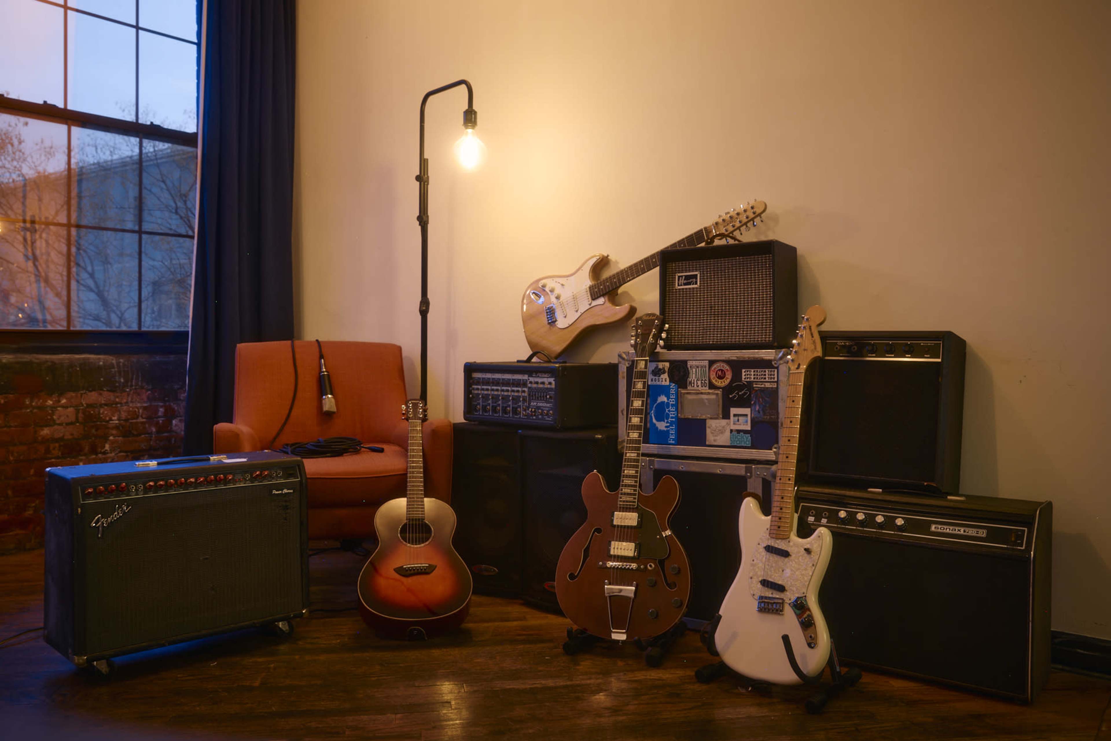 The scene features a collection of musical instruments, including electric and acoustic guitars, amplifiers, and a lamp, arranged in a cozy room with a large window.