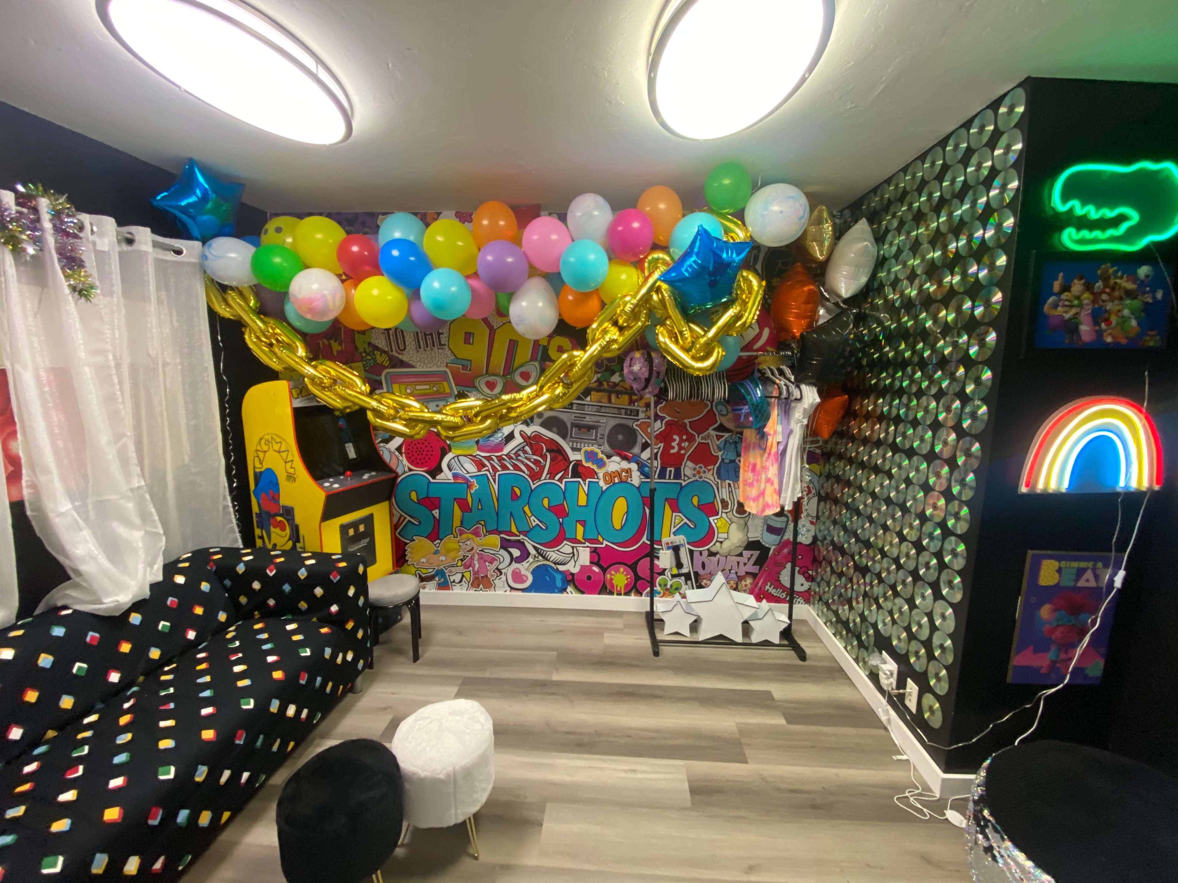 The image shows a vibrant party room adorned with colorful balloons, a large banner reading "STARSHOTS," and various decorations on the walls, including sequined panels and a neon rainbow light.