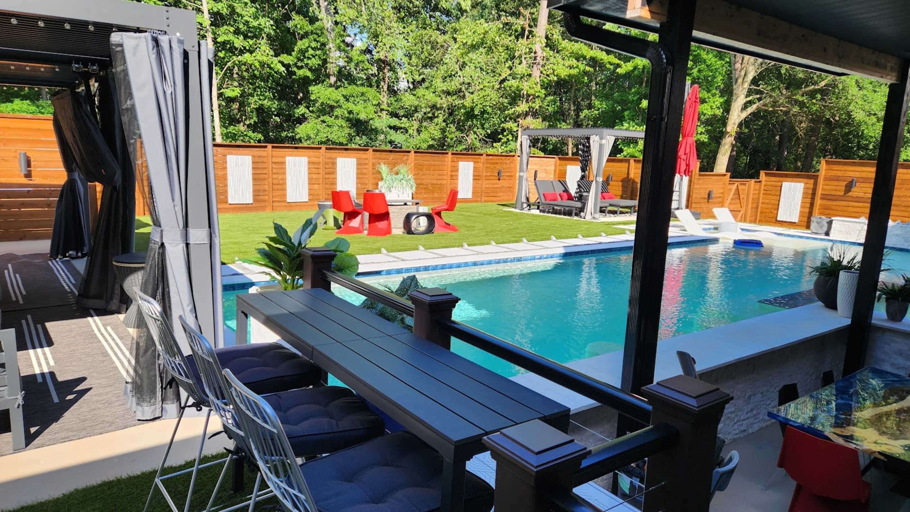 The image shows a backyard with a swimming pool surrounded by lounge chairs, patio furniture, and greenery.