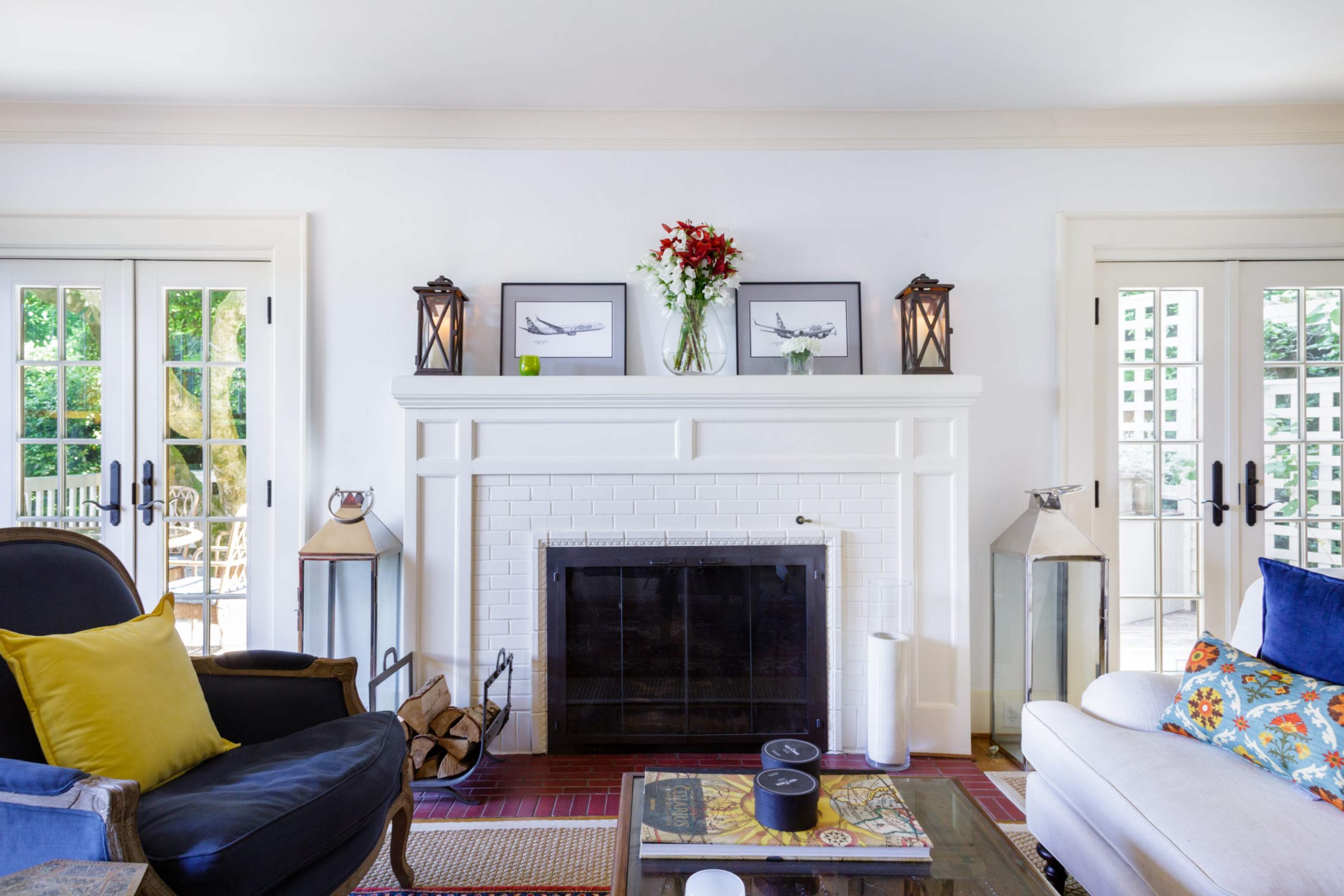 A living room with a white brick fireplace flanked by decorative lanterns and a vase of flowers, alongside two seating areas and french doors leading outside.