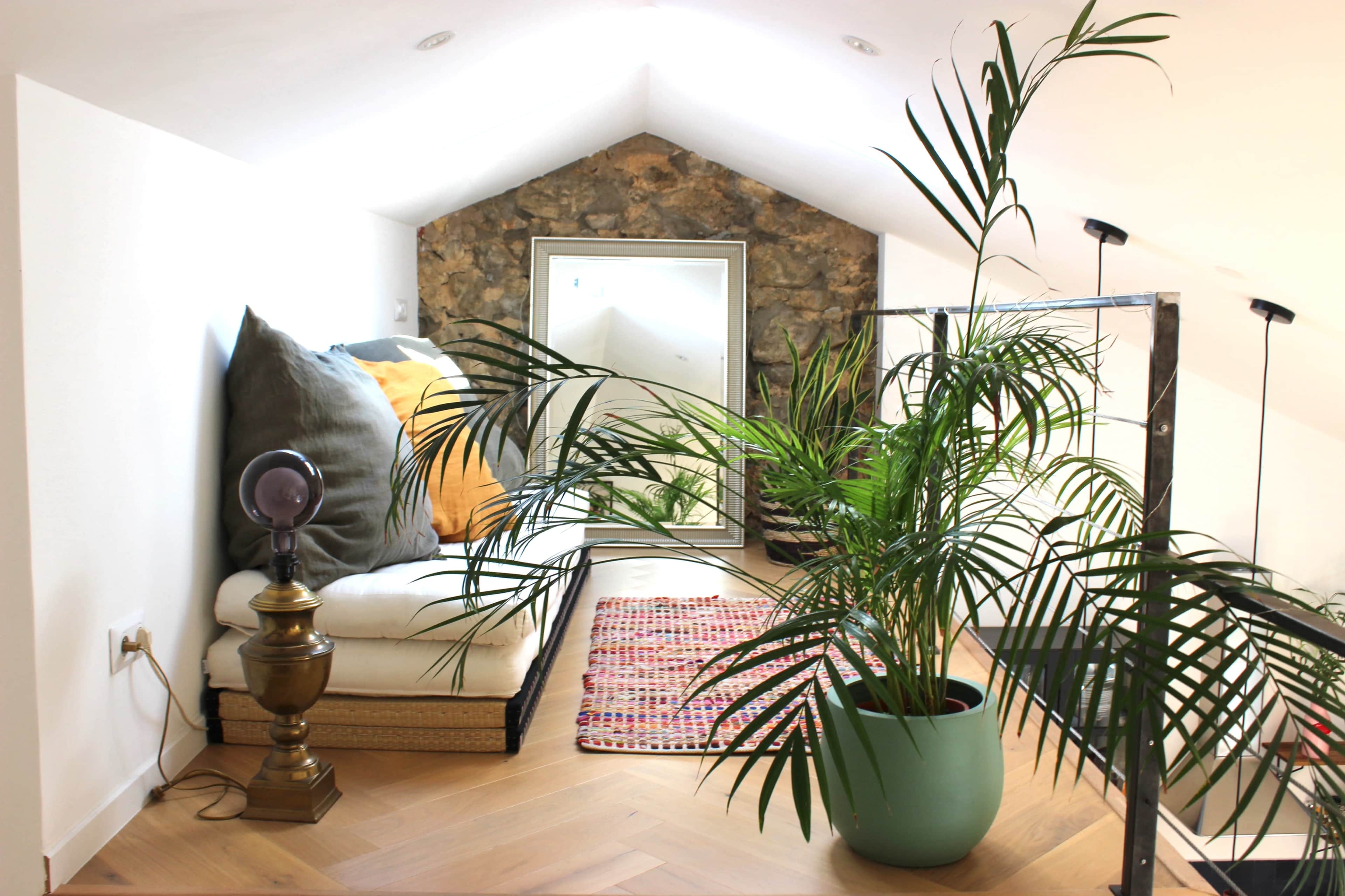 A cozy loft space features a seating area with large cushions, a mirror, a stone accent wall, and several potted plants.