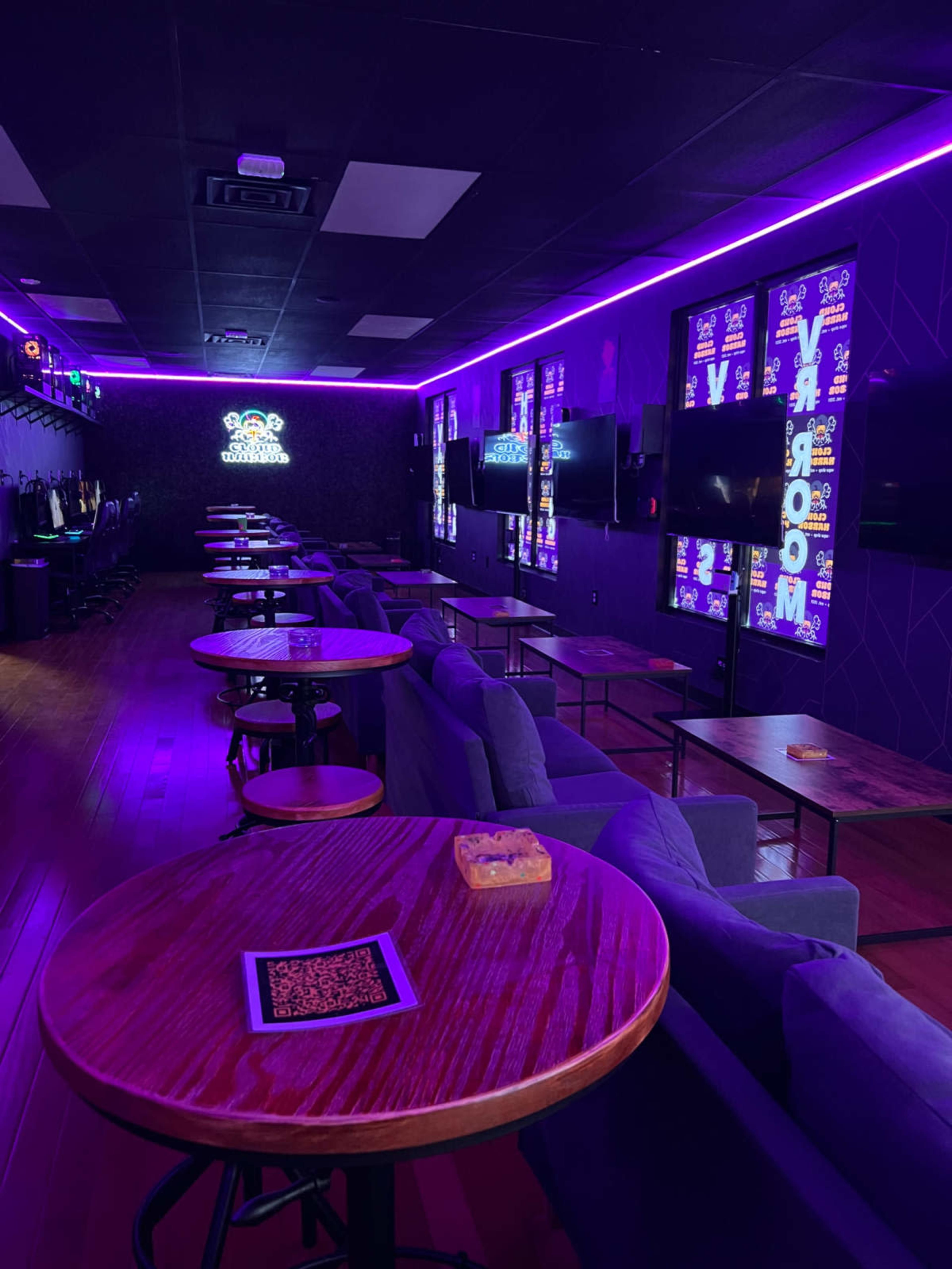 The image shows a dimly lit lounge area with tables and blue chairs, featuring purple LED lighting along the walls and multiple screens displaying graphics.