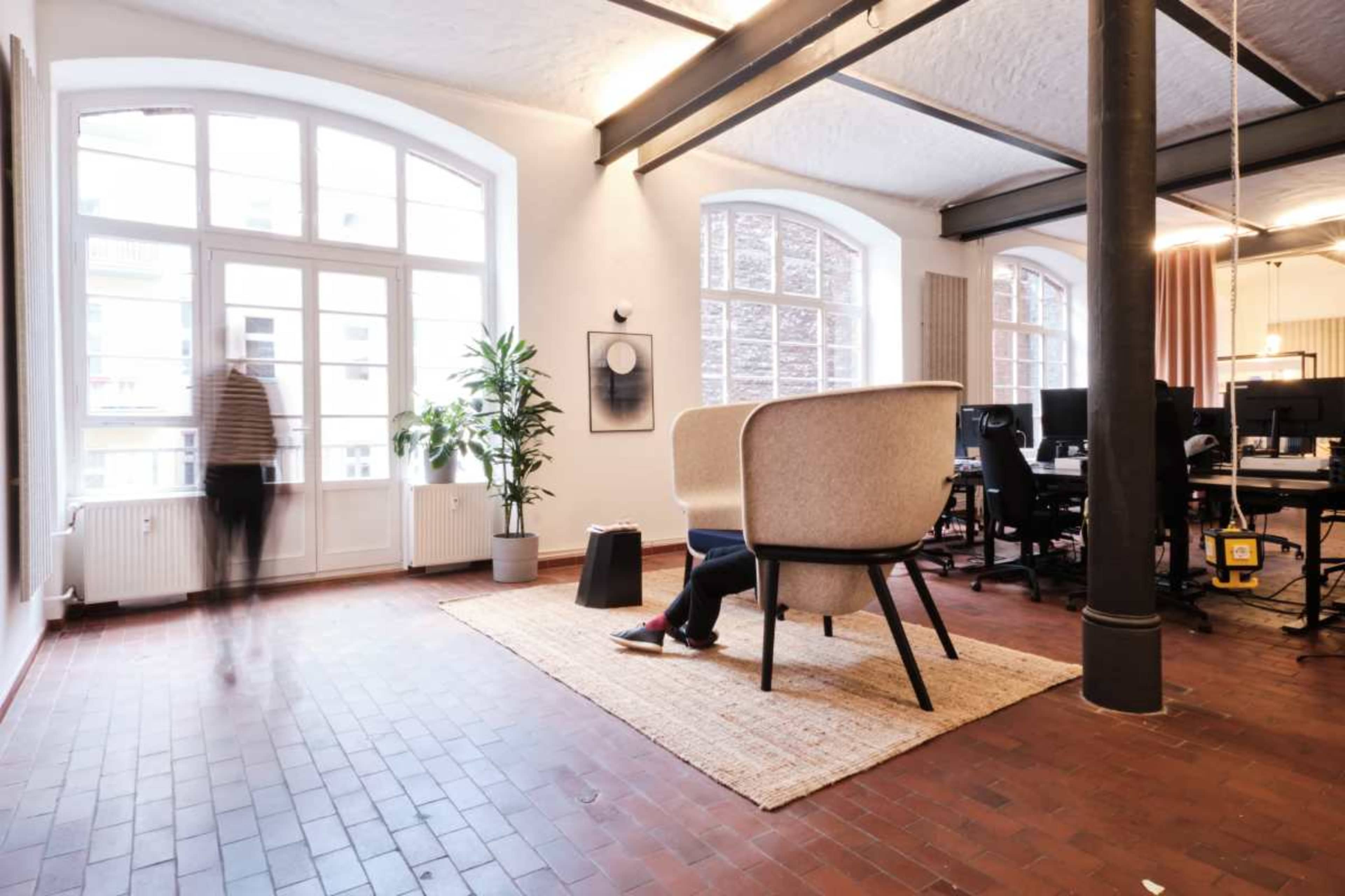 A spacious office interior features a large window, modern seating arrangements, a plant, and workstations in the background.