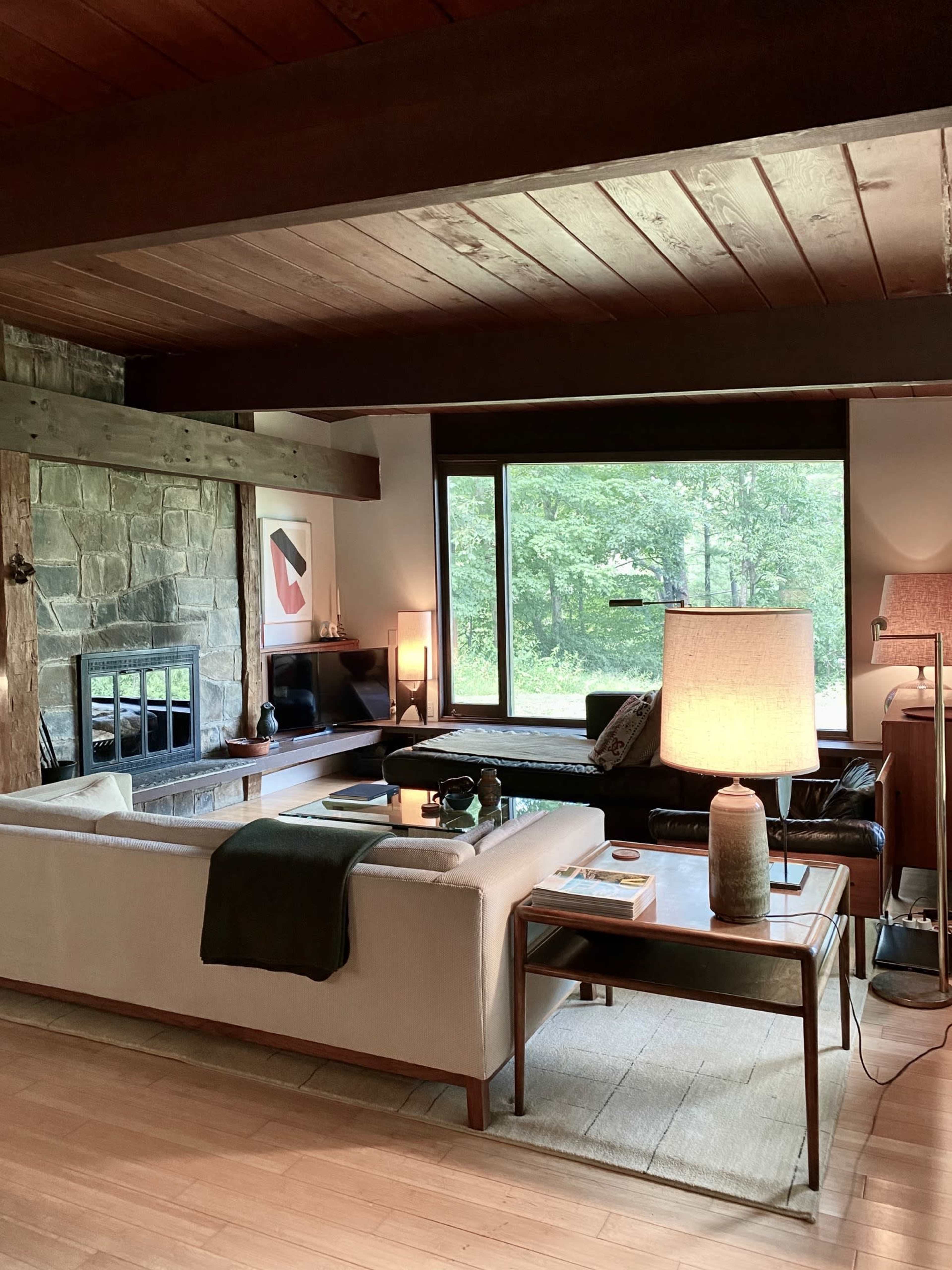 The image shows a modern living room featuring a stone fireplace, large windows overlooking greenery, a sectional sofa, and a wooden coffee table with decorative items.