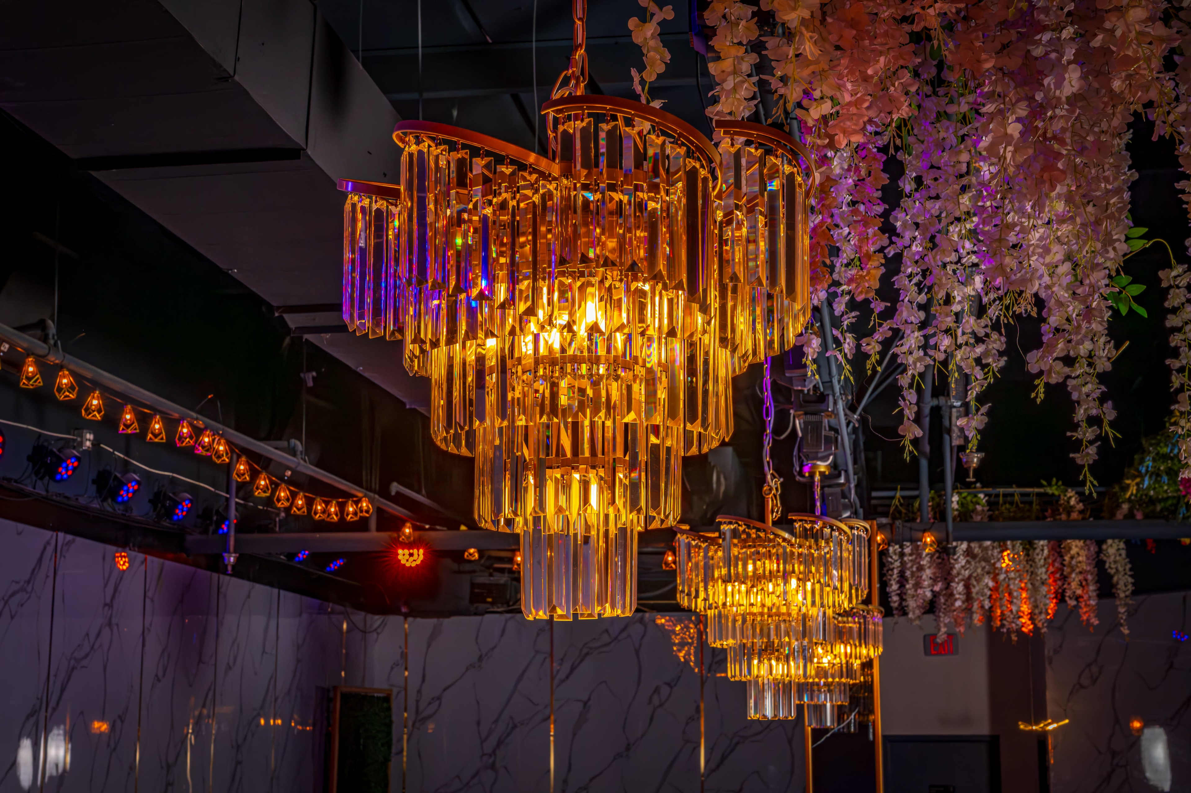 Three elegant chandeliers made of glass tubes hang from the ceiling, illuminated by warm lights, amidst decorative floral arrangements.