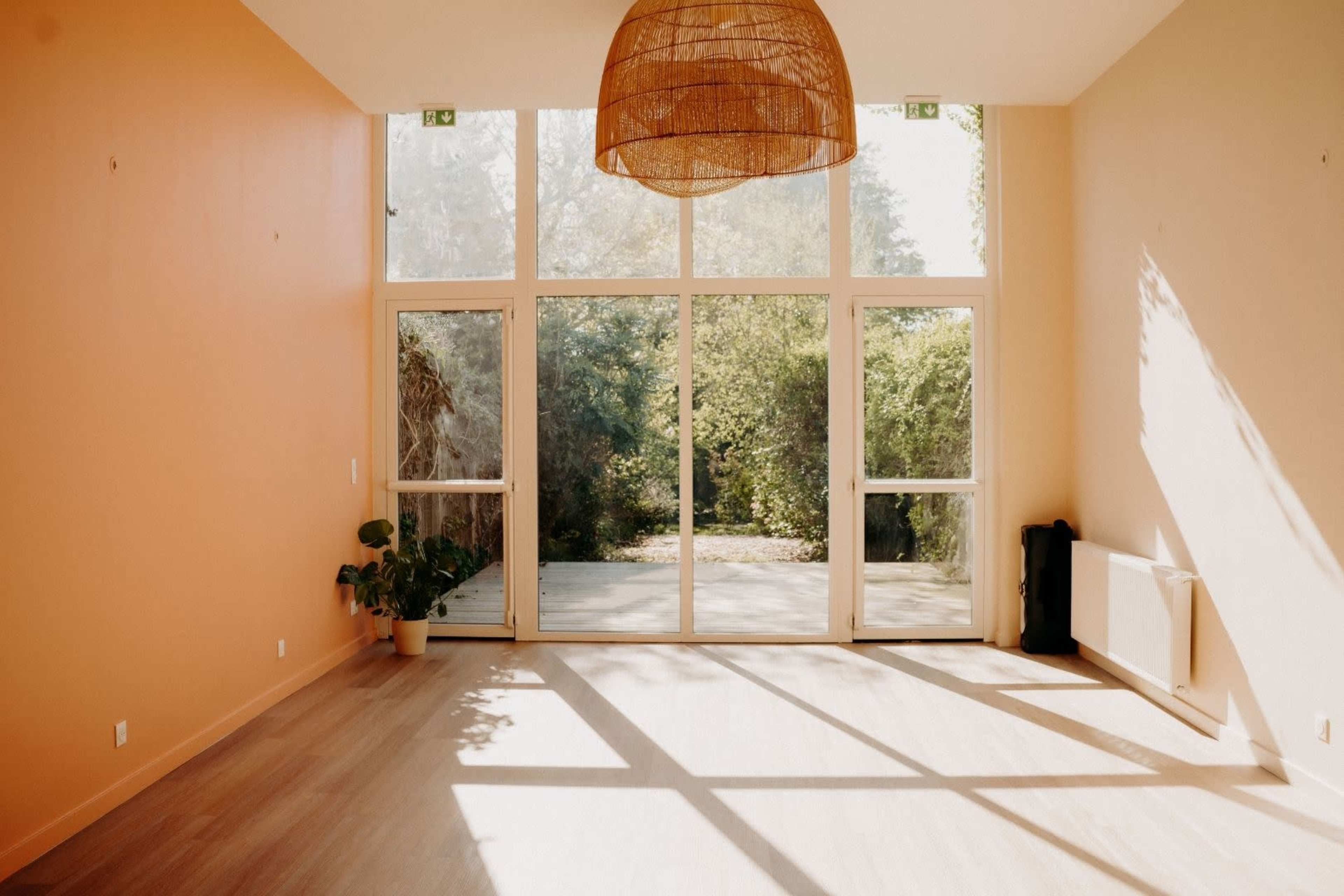 The image shows a bright, empty room with orange walls and large glass windows that overlook a green outdoor space.