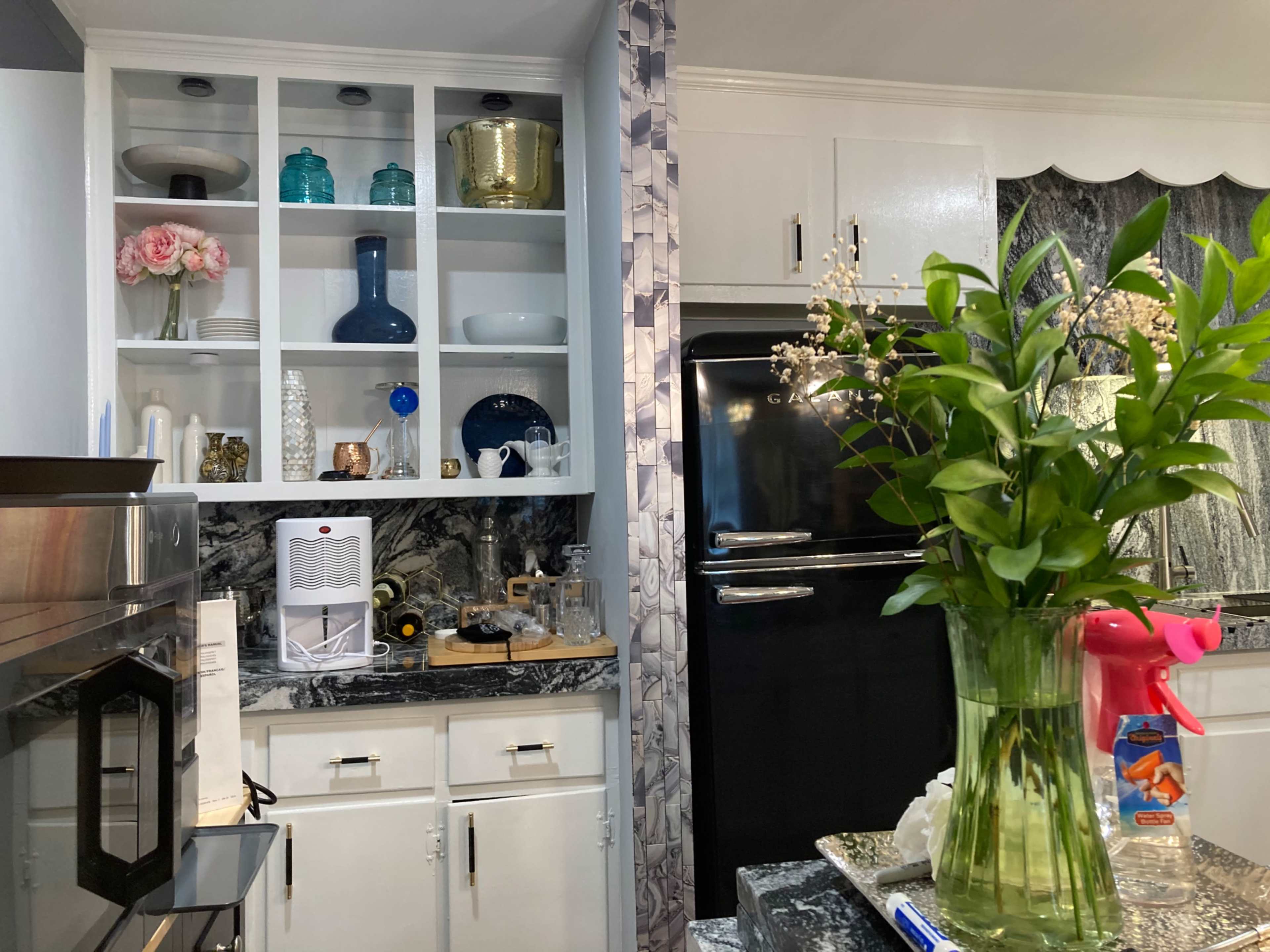 A kitchen with upper shelves displaying decorative items and tableware, alongside a black refrigerator and a counter with cleaning supplies and a vase of flowers.