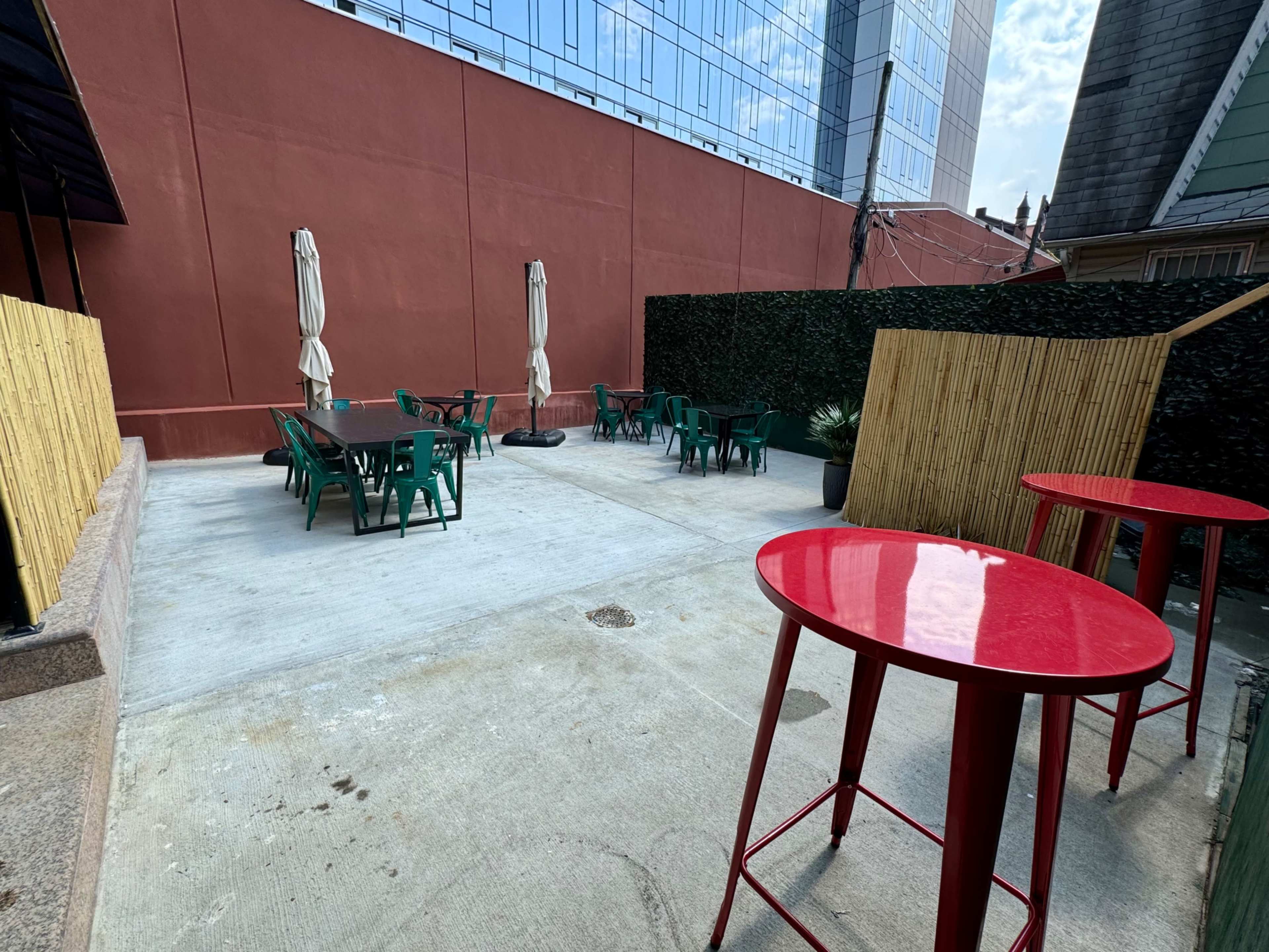 The outdoor patio features several green chairs and tables, along with two red high tables, surrounded by a concrete floor and a bamboo fence.