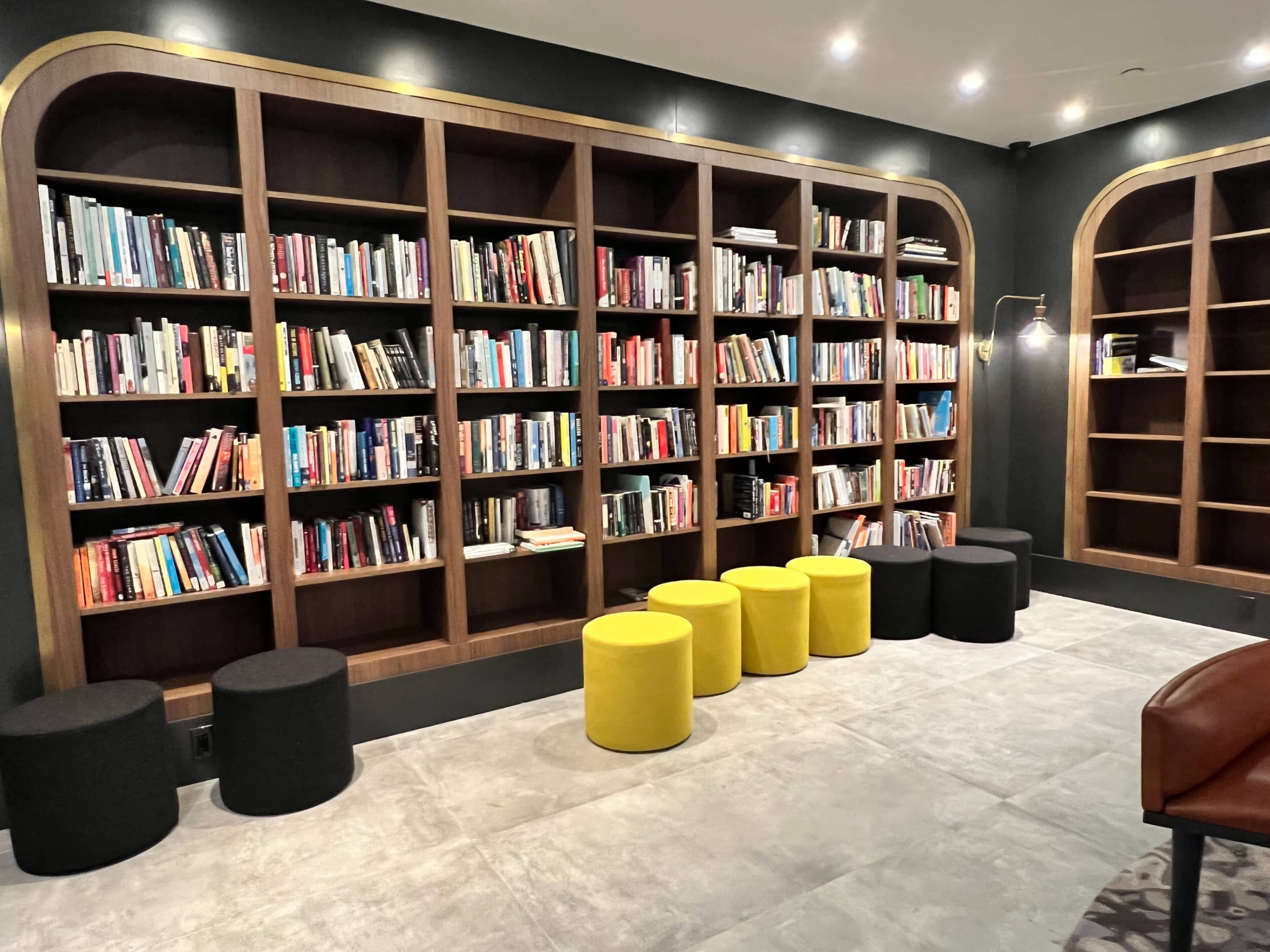 The image shows a modern room with a large bookshelf filled with books and a row of circular stools in black and yellow.